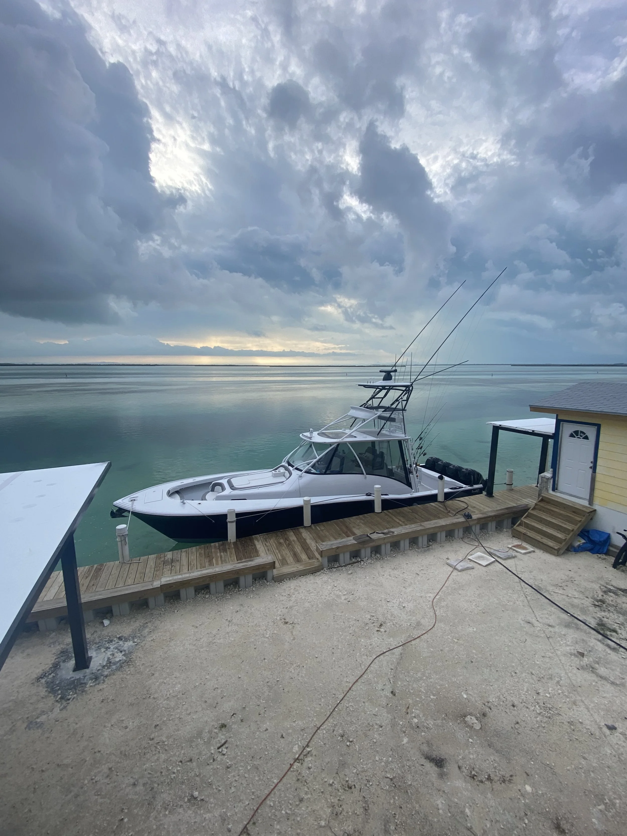 A boat docked at a wooden pier by a small yellow building on a sandy area with clouds overhead and water in the background.