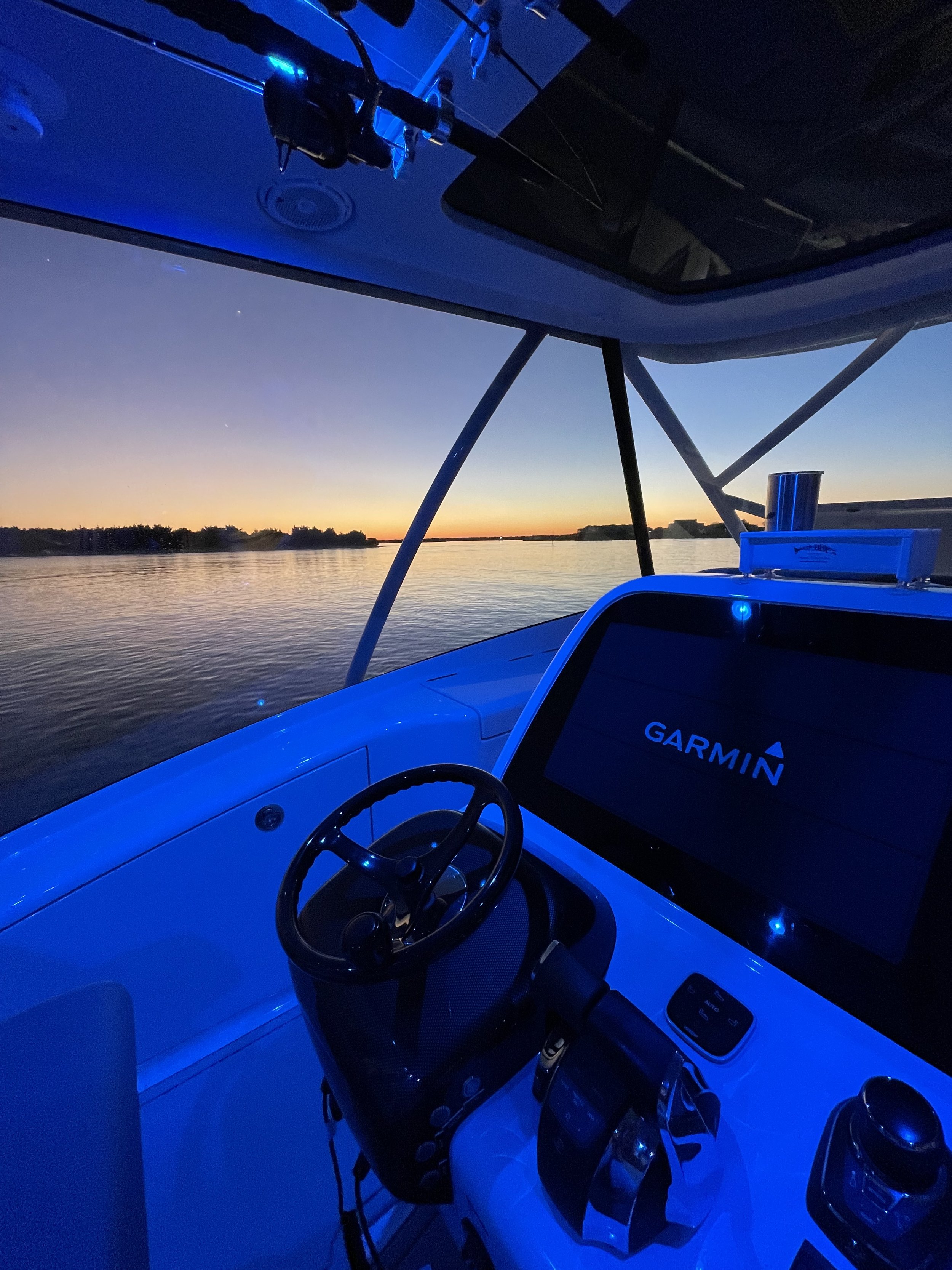 Inside a boat at sunset with a view of the calm water and distant shoreline, featuring a steering wheel, Garmin navigation screen, and electronic devices illuminated by blue lighting.