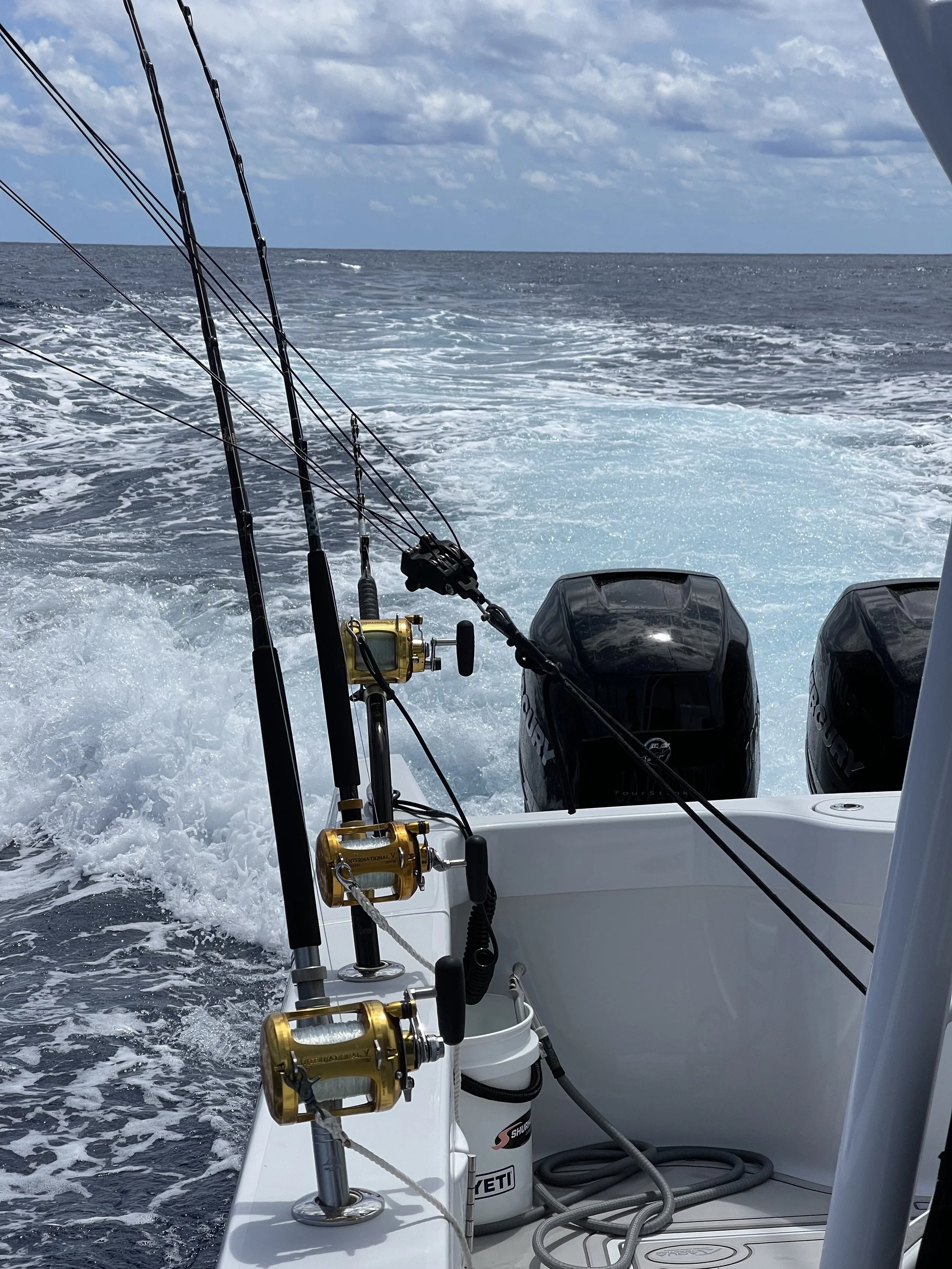View from a boat showing three fishing rods mounted at the stern, with the boat moving through the water leaving a wake behind, and the ocean with clouds overhead.