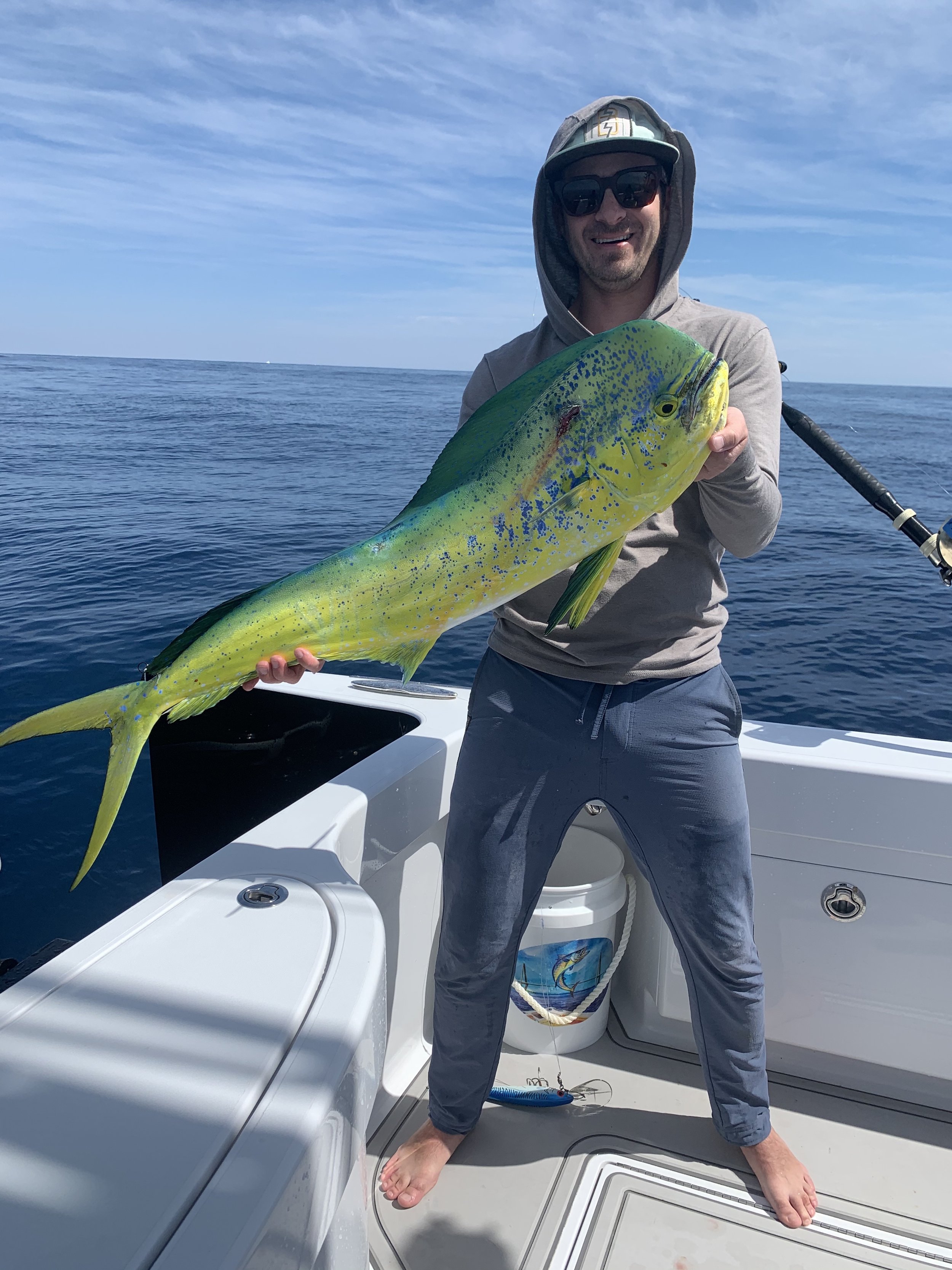A man on a boat holding a large, colorful fish with yellow, green, and blue markings against a calm ocean and blue sky background.