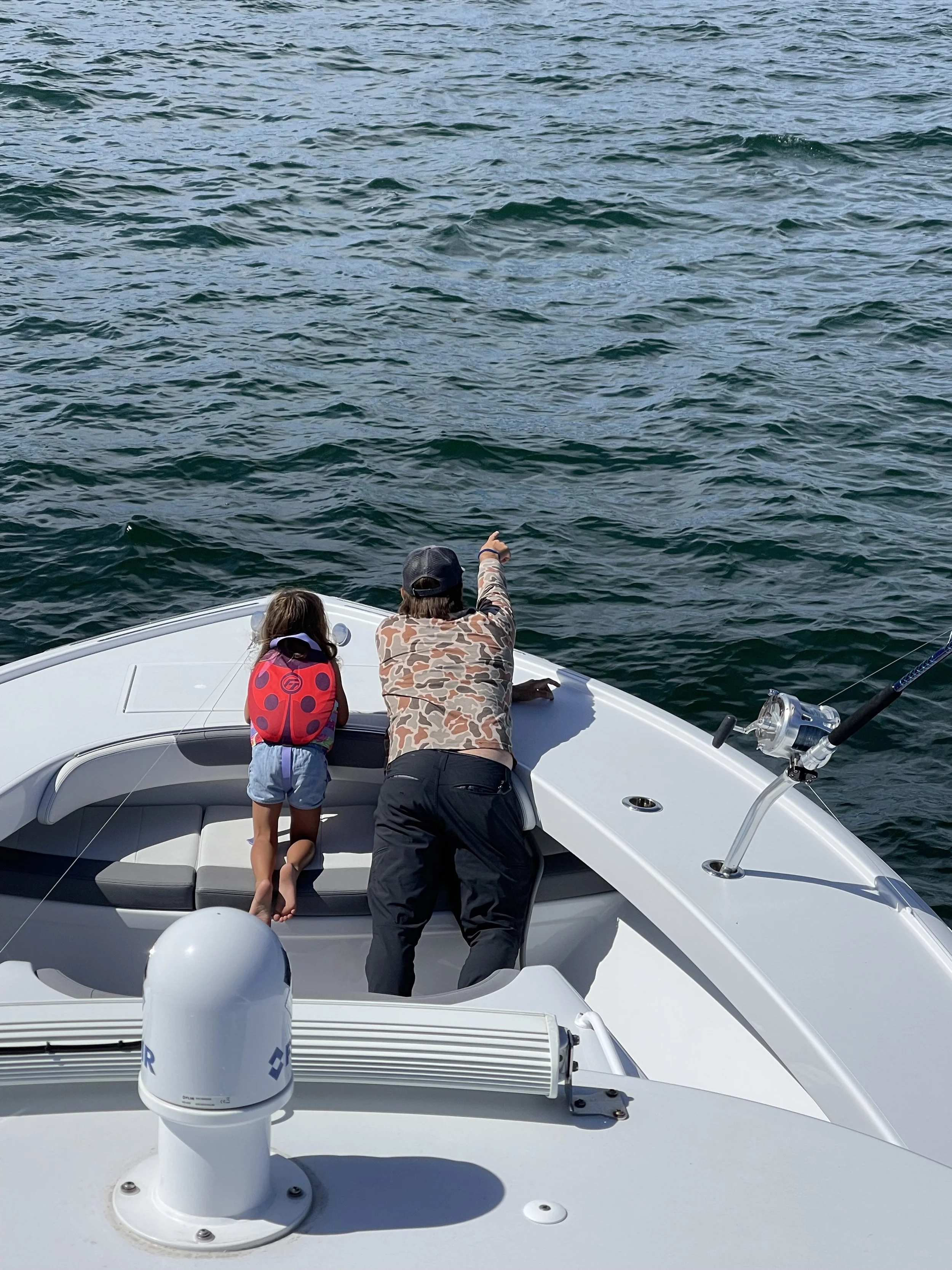A man and a young girl on a white boat, with the girl wearing a red life jacket, are looking and pointing towards the water.