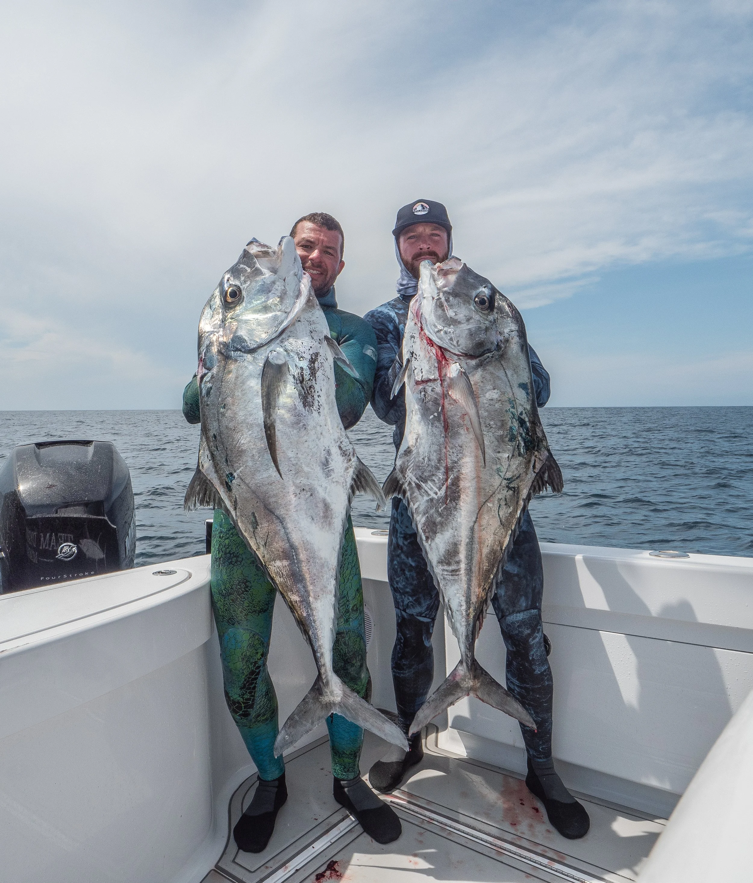 Two men on a boat holding large fish they caught, standing on the deck of the boat with ocean and sky in the background.