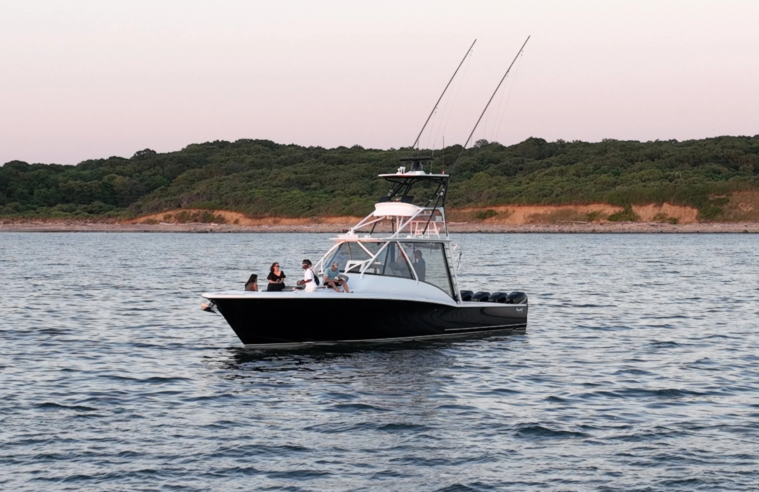 A group of five people on a black and white cruiser boat on calm water with a wooded shoreline in the background during sunset.