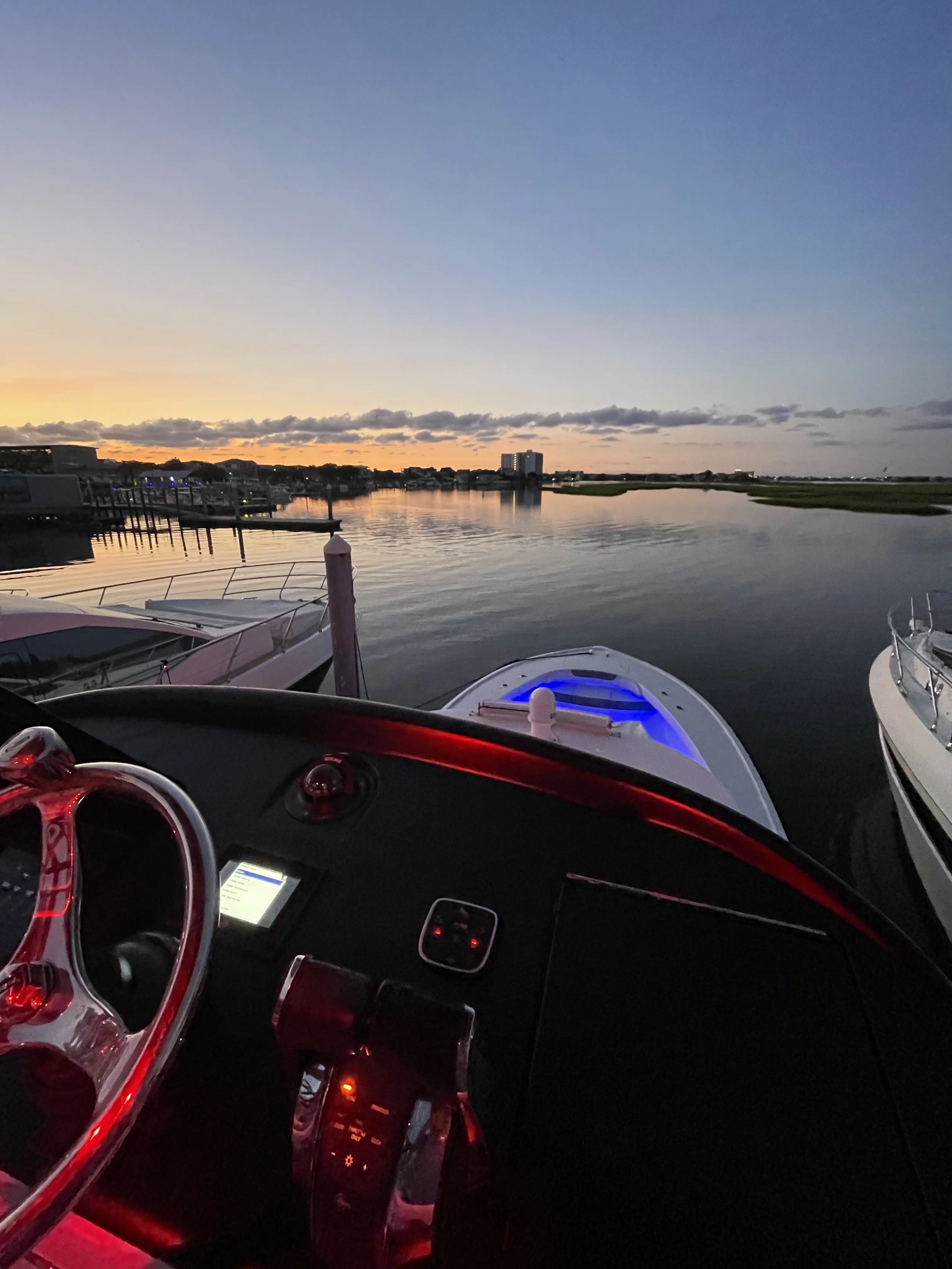 View from the cockpit of a boat at sunset, showing calm water, other boats docked nearby, a distant cityscape, and a colorful sky.