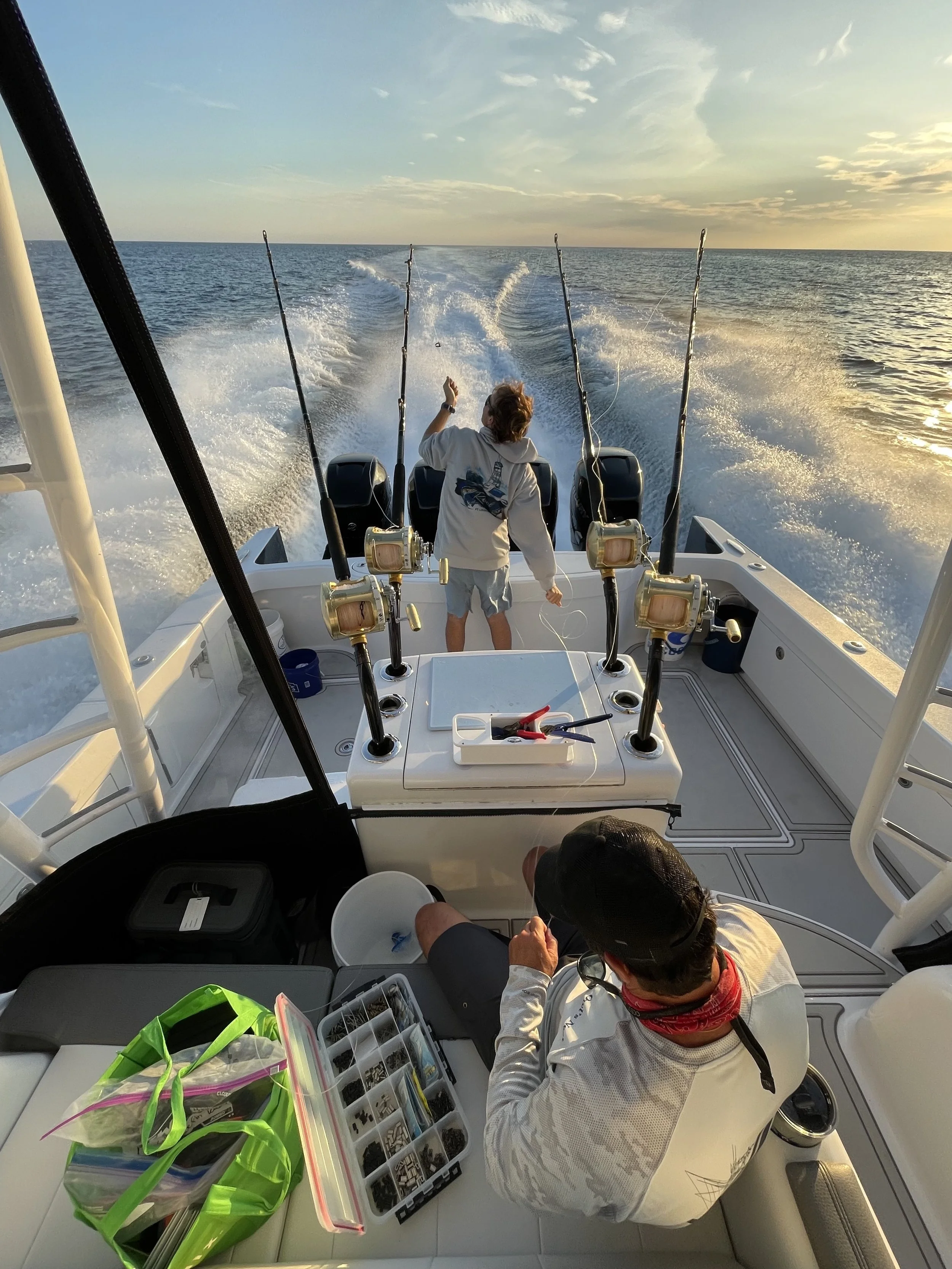 A person fishing on a boat at sunset with four fishing rods, with another person sitting nearby in a white shirt, cap, and red bandana, surrounded by fishing tackle and gear.