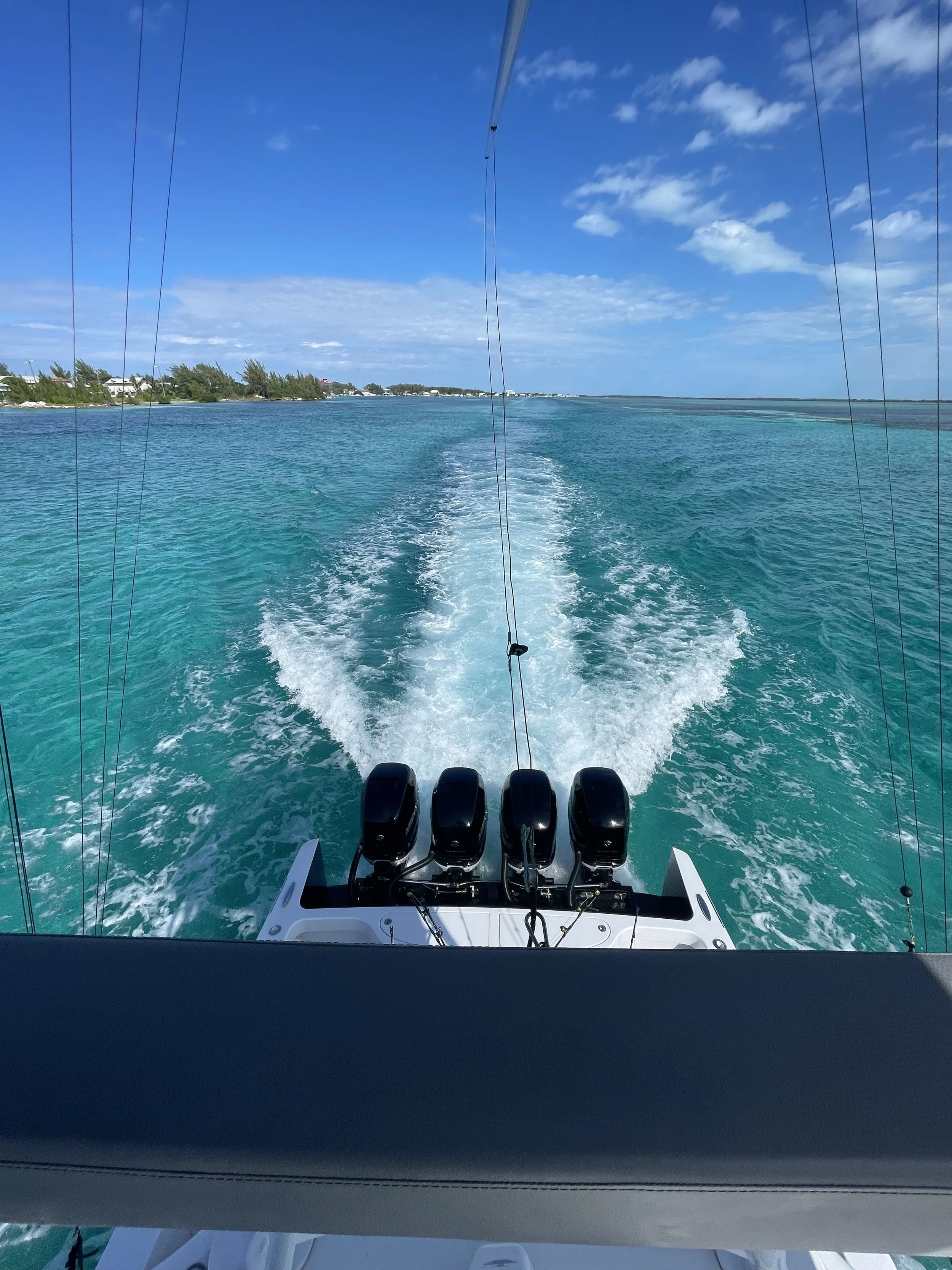 View from the back of a boat showing the engine and wake in the turquoise ocean, with a coastline and partly cloudy sky in the distance.