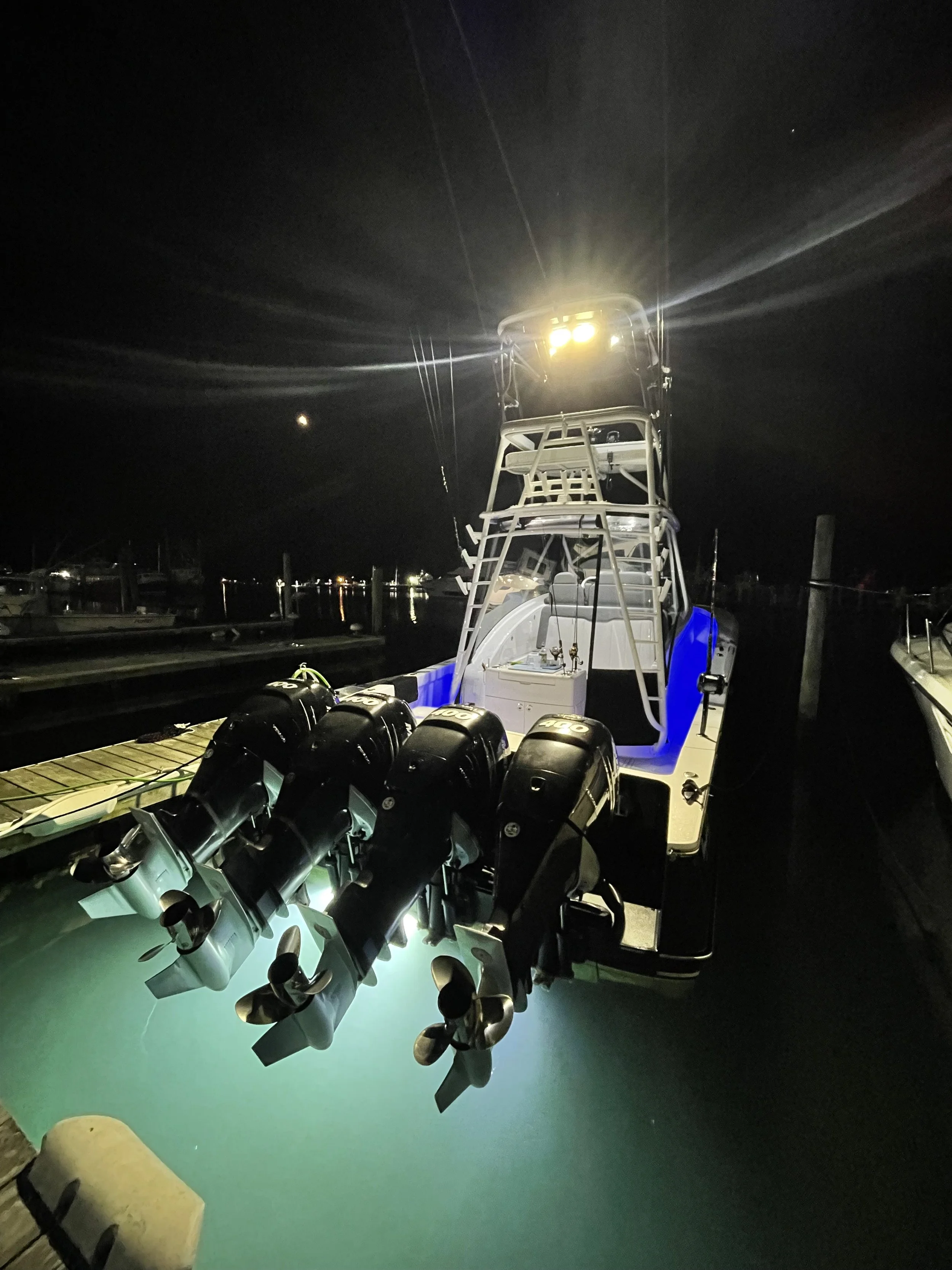 Night shot of a fishing boat docked at a marina, equipped with four outboard motors, bright lights at the top illuminating the boat, and a blue cushion on the deck, with a dark sky and faint reflections on the water.