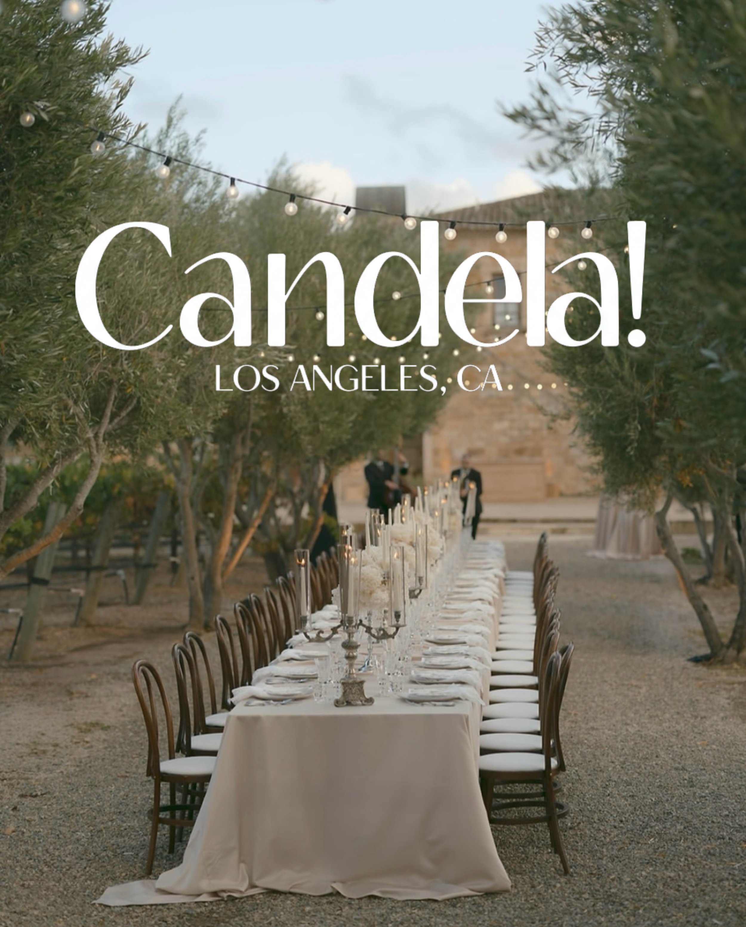 An outdoor dining table decorated with white tablecloths, candle holders, and candles, set in a garden with trees and string lights overhead, for a special event in Los Angeles, California.