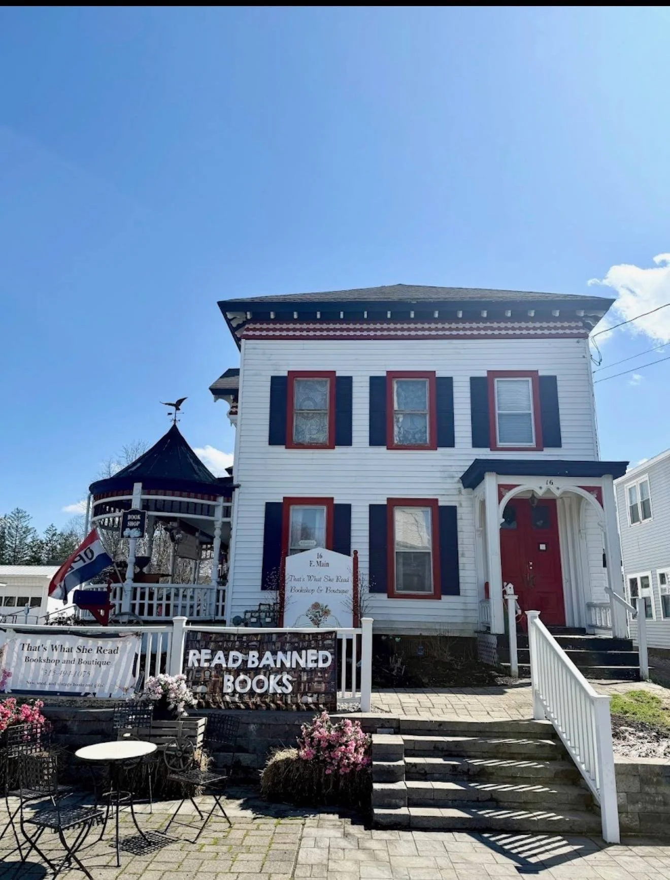 A white, three-story house with red and black trim, featuring a small porch and a turret with a weathervane on top. There are signs in front that read ‘READ BANNED BOOKS’ and That's What She Read Bookshop and Boutique. The area has outdoor seating with chairs and flowers, and a white picket fence surrounds the front yard.