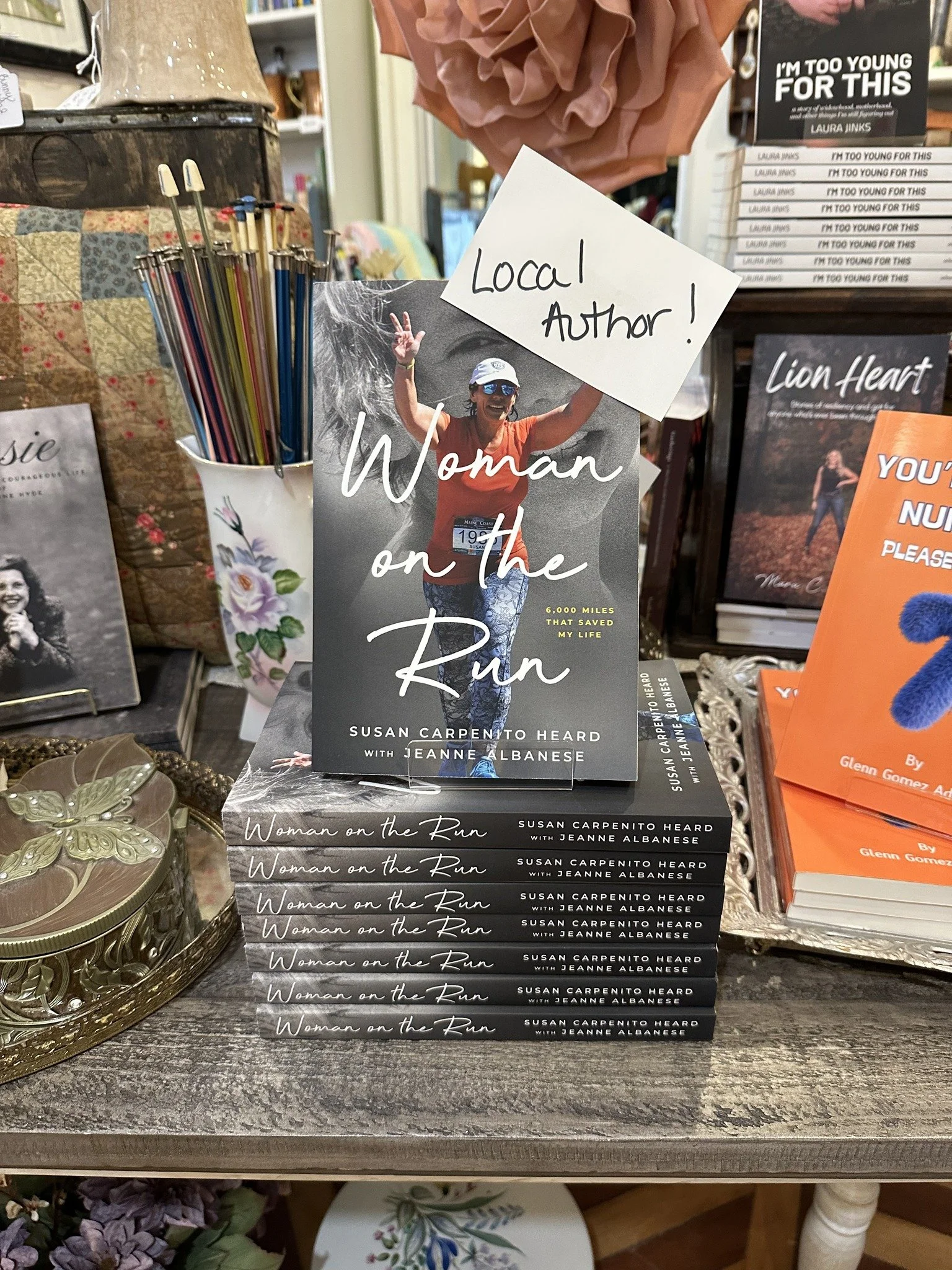 The image shows a display of a book titled "Woman on the Run" by Jeanne Albanese. The book cover features a woman running outdoors, wearing a hat and sunglasses, raising her hand in a victory sign. There is a handwritten sign that says "Local Author!" placed on the book. The background includes other books, decorative items, and a pen holder with colorful pens.