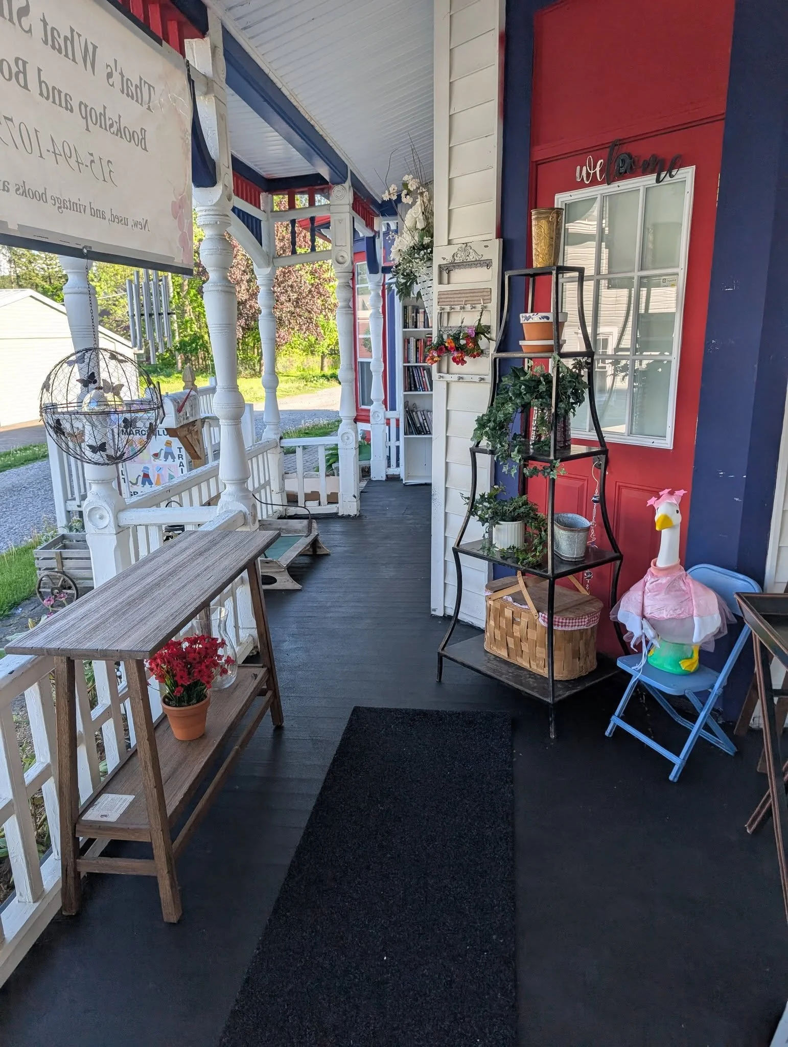 A front porch with a black welcome mat, white porch railing, and a white lattice. Decor includes a black wire hanging orb with butterflies, a wooden table with a potted red flower, a white bookshelf with flowers and books, and a black metal plant stand with greenery. A pink flamingo lawn ornament and a blue folding chair are nearby. The porch has red and blue walls, a window with multiple panes, and a sign reading 'we belong' above the window. Outside trees and a house are visible in the background.