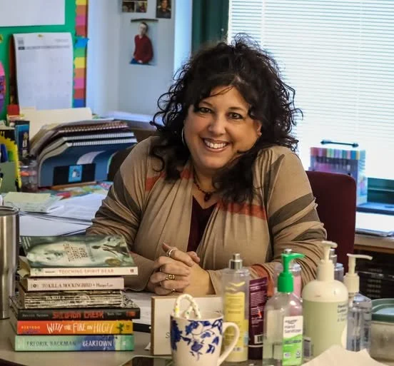 A woman with curly dark hair smiling at her workspace, surrounded by books, papers, and hand sanitizer bottles, in a colorful classroom or office setting with a window and various decorations in the background.