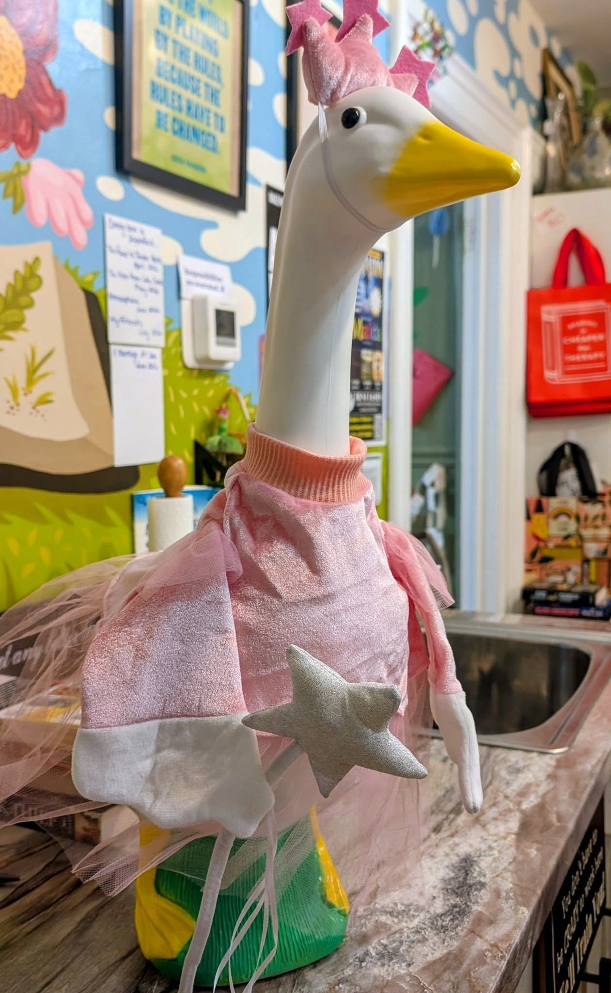 A decorative large ceramic or plastic goose figure dressed in a pink dress with white stars and a pink felt crown with stars on its head. It appears to be part of a display and is placed on a table in a room with colorful wall art and various household items in the background.