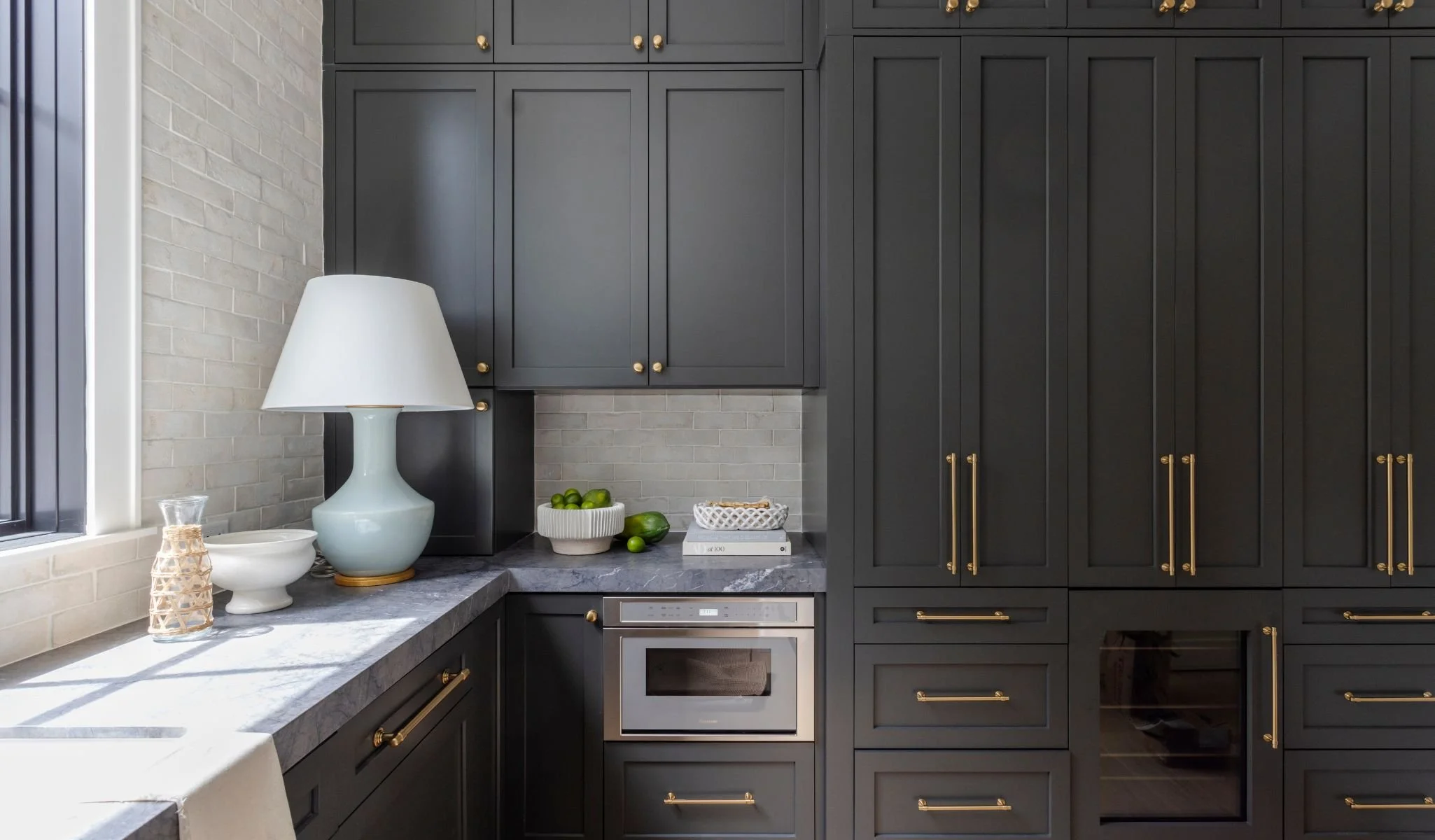 Kitchen with dark gray cabinets with gold handles, a marble countertop, a window with sunlight, a white table lamp, decorative bowls, and limes on the counter.