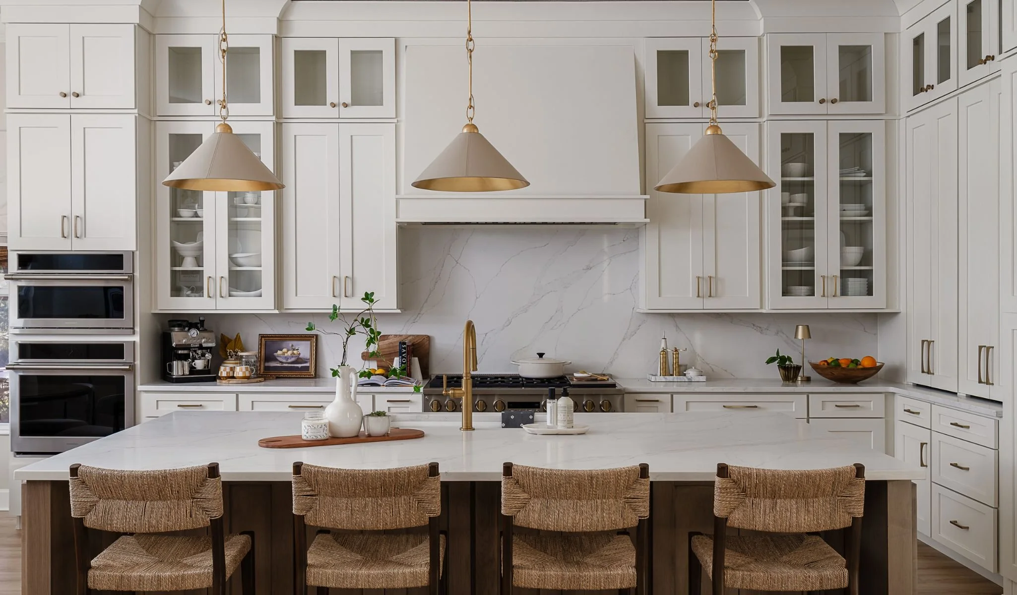 A modern white kitchen with a large island, gold accents, hanging pendant lights, and glass-front cabinets filled with dishes and glassware.