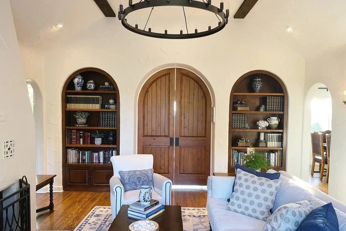 Living room with white sofa and armchair, decorative pillows, wooden coffee table with books, custom made bookshelf with decorative items and books, large wooden double door, and a chandelier hanging from the ceiling.