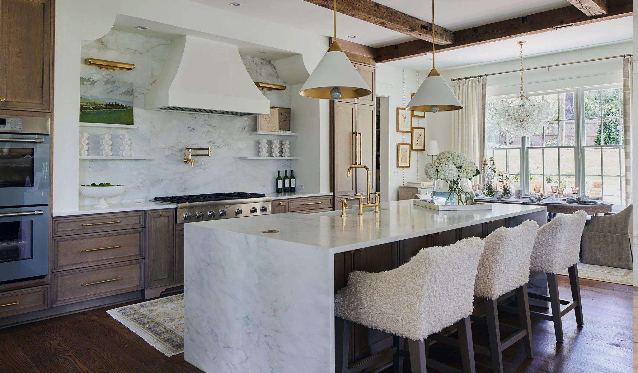 A modern kitchen with a large white marble island, gold fixtures, wooden cabinets, and a dining area with a table set for a meal near large windows with a view of a backyard.