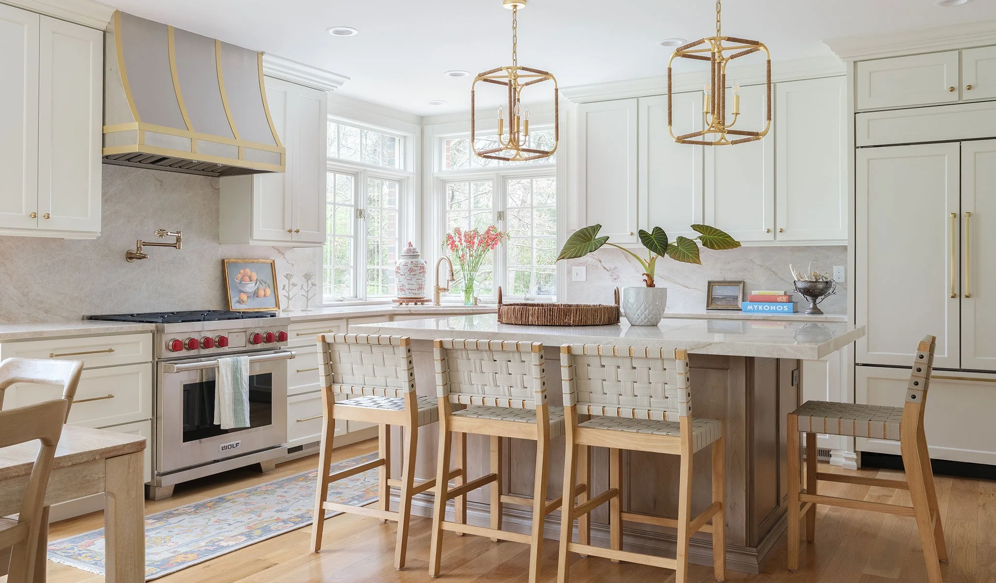 Bright kitchen with white cabinets, gold hardware, a marble island with woven chairs, plants, flowers, and natural light coming through large windows.
