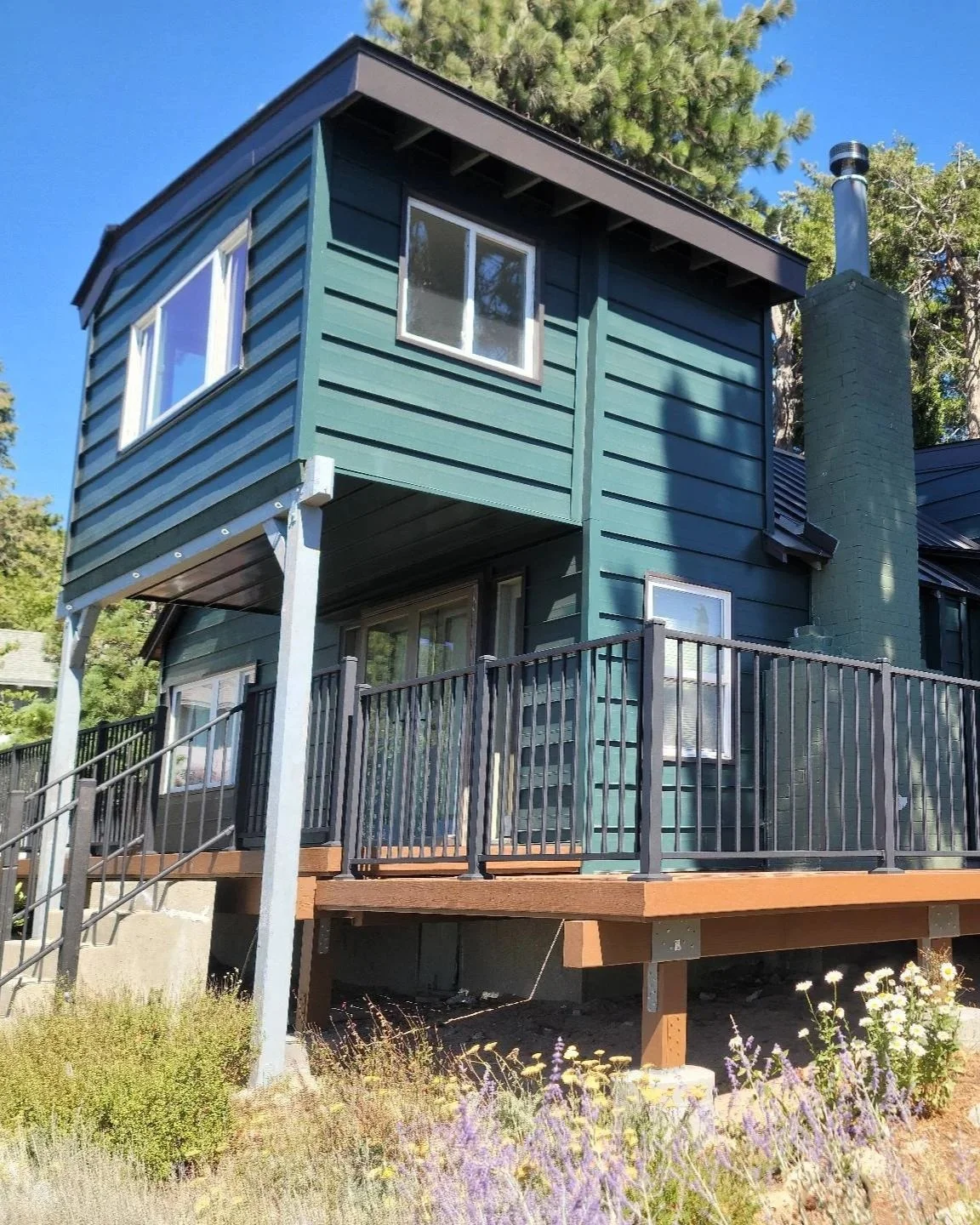 A house under construction on a sloped lot with a large wooden deck, surrounded by trees, and workers installing the foundation.