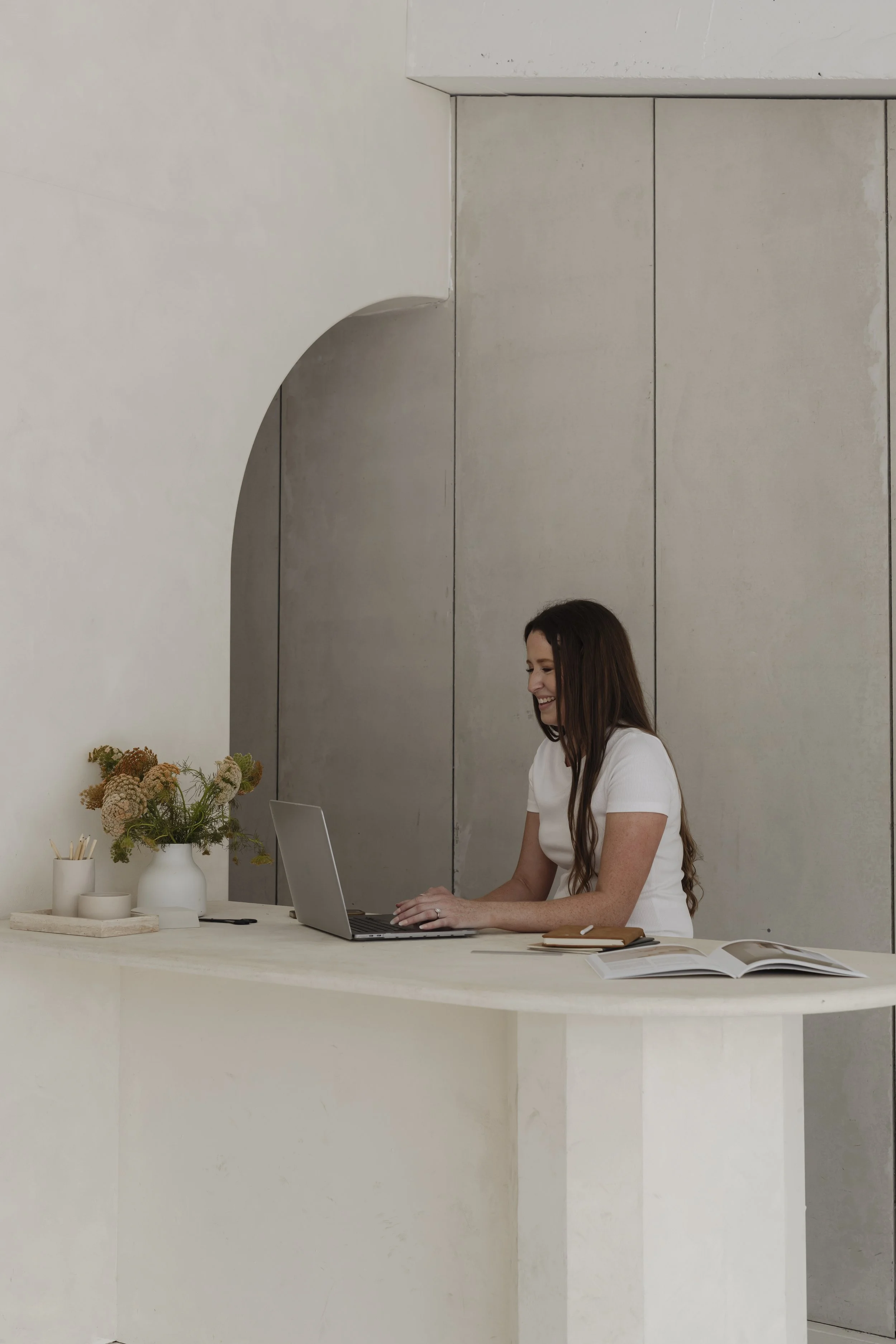 A woman with long brown hair sits at a white desk, smiling, working on a laptop, with a vase of flowers, notebooks, and an open magazine on the desk, against a minimalist gray wall.