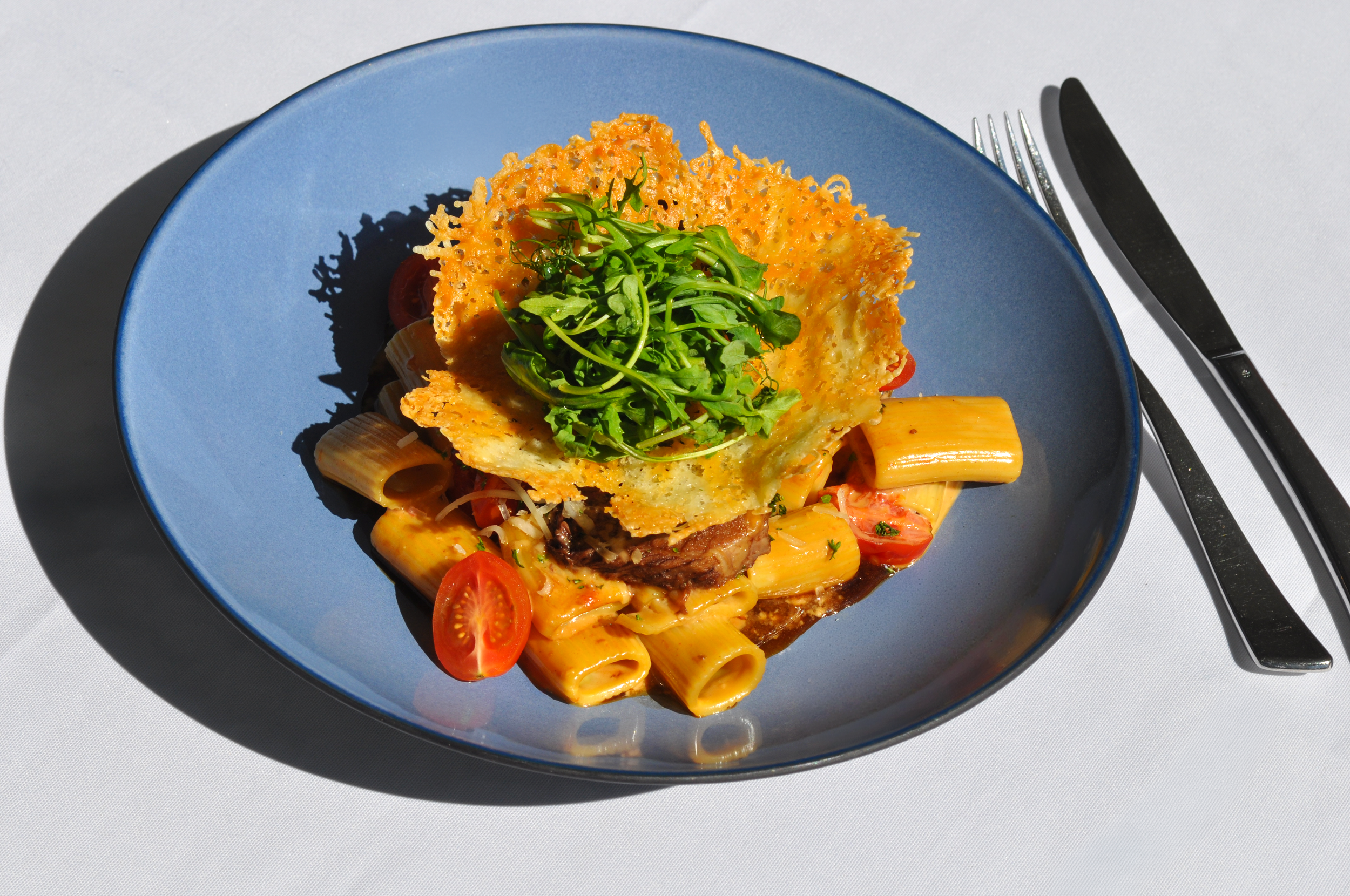 Gourmet rigatoni pasta with braised meat, cherry tomatoes, greens, and a crispy Parmesan tuile on a blue plate