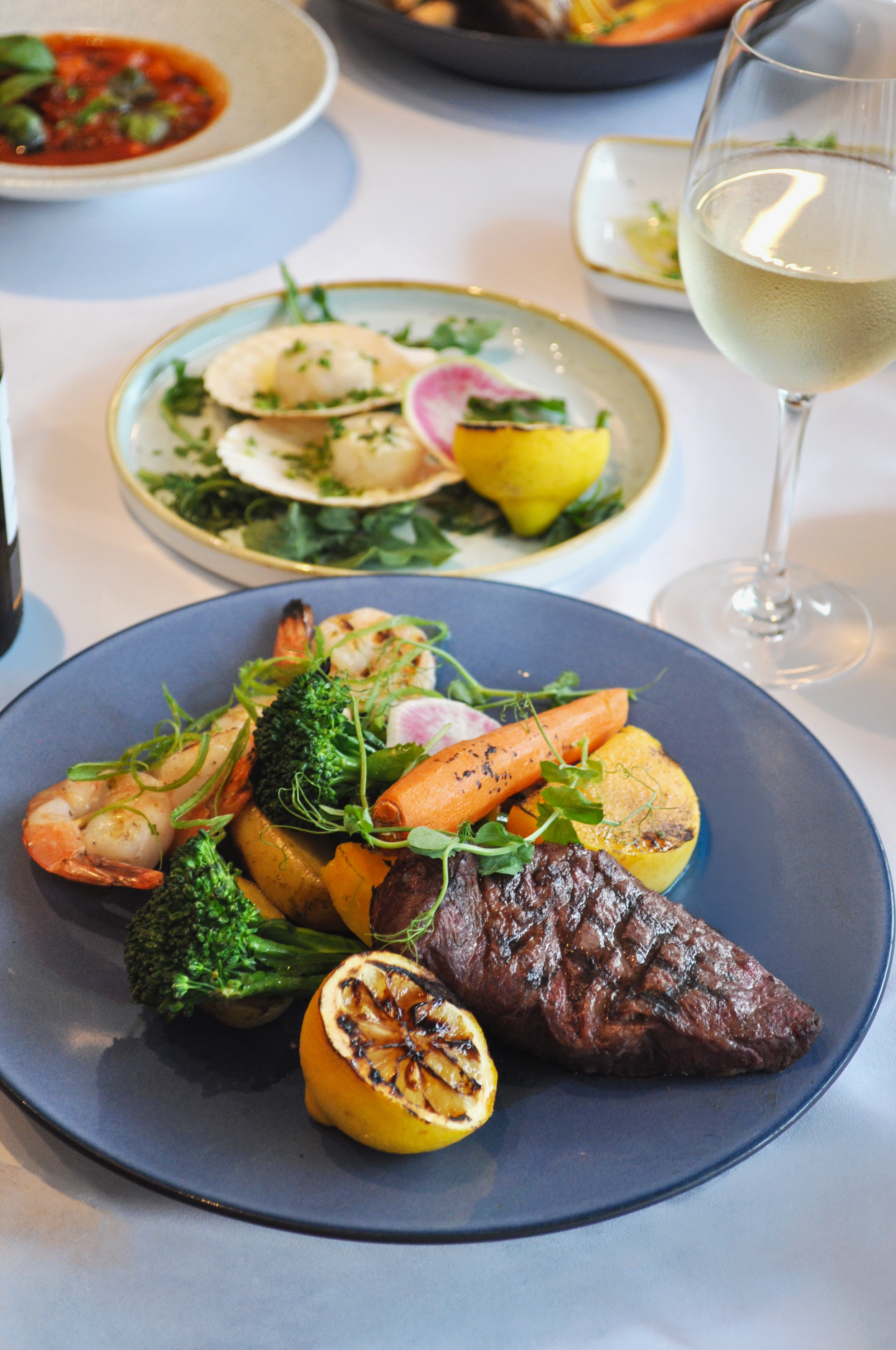 Elegant restaurant spread featuring a grilled steak with shrimp, roasted vegetables, broccolini, and grilled lemon, alongside seafood dishes and white wine.