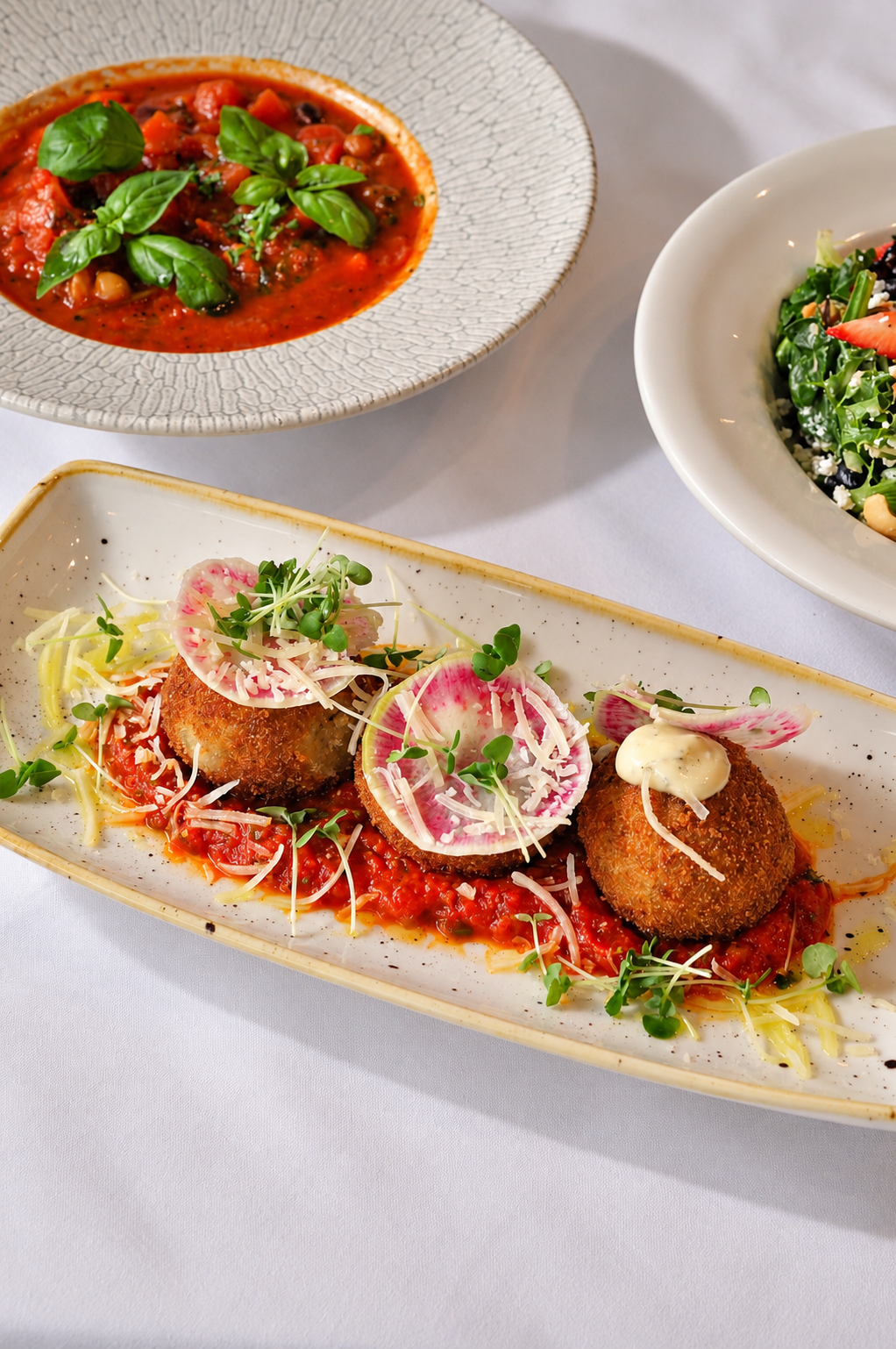 Restaurant spread featuring breaded croquettes with radish slices and microgreens over tomato sauce, tomato basil soup, and a green salad