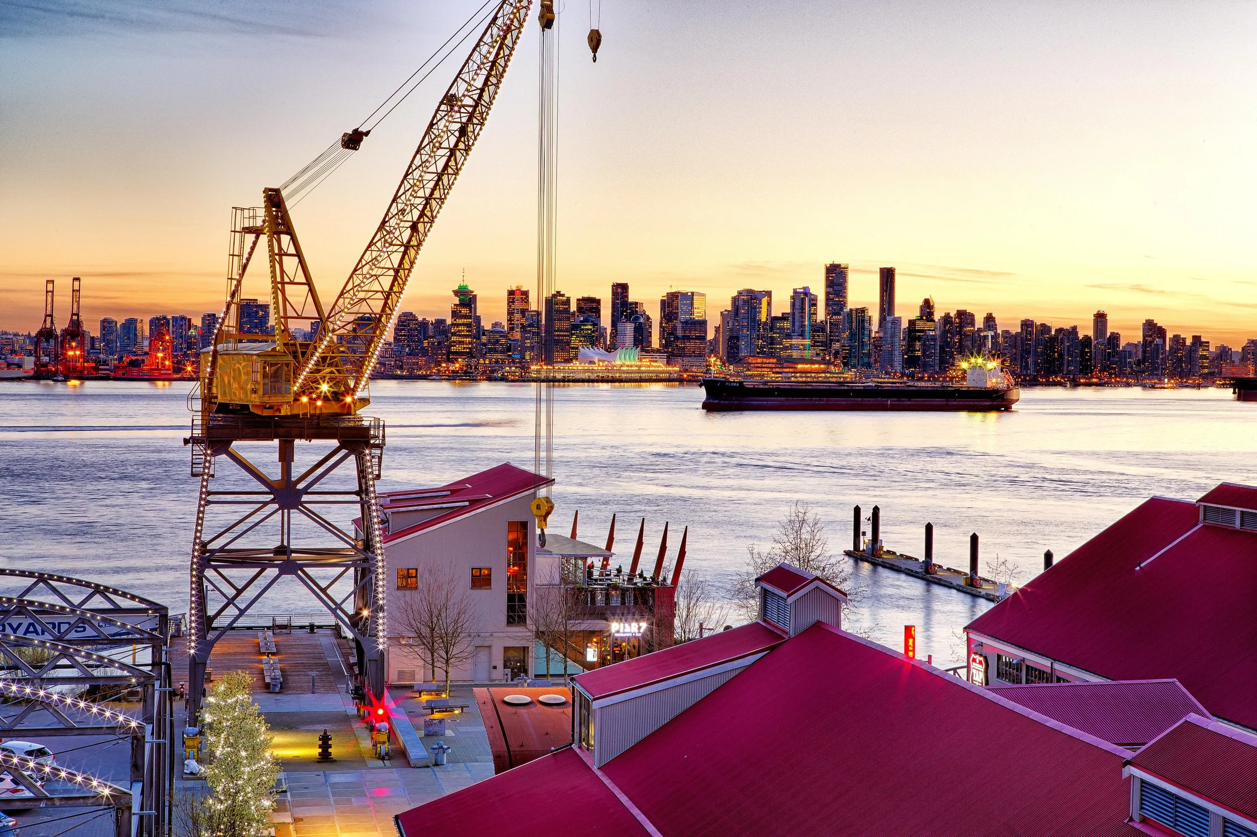 Vancouver skyline at sunset viewed from Granville Island, with a waterfront construction crane, cargo ship, and illuminated high-rise buildings.