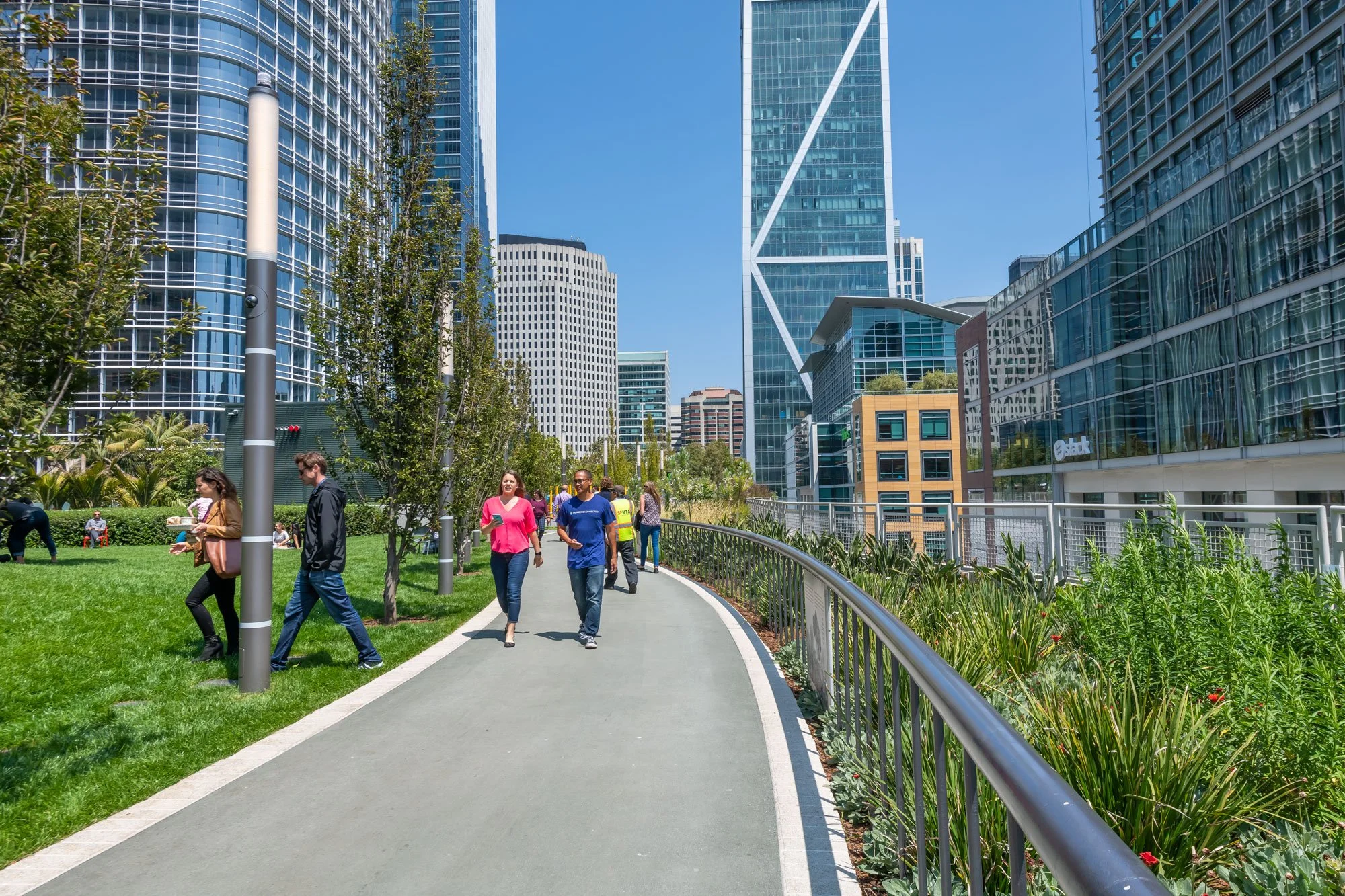 People walking on a paved sidewalk beside a green lawn with trees, surrounded by modern office buildings under a bright blue sky.