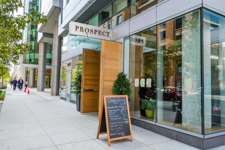 Modern city street scene featuring a building with large glass windows and a sign that reads "PROSPECT". There is an open wooden door, a blackboard sign with handwritten menu, and some potted plants outside. Pedestrians walk along the sidewalk in the
