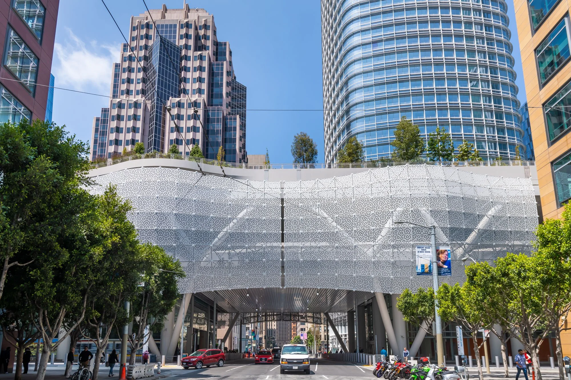 City street with modern buildings, trees, and a unique architectural structure resembling a giant, perforated white shell with a metallic underside, cars, bikes, and pedestrians.