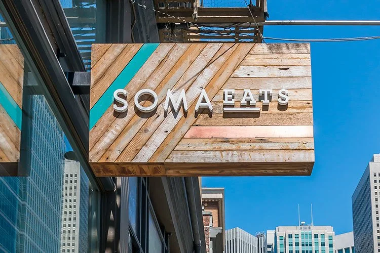 Wooden sign with the text 'SOMA EATS' hanging outside a building in an urban area with tall skyscrapers and a clear blue sky.