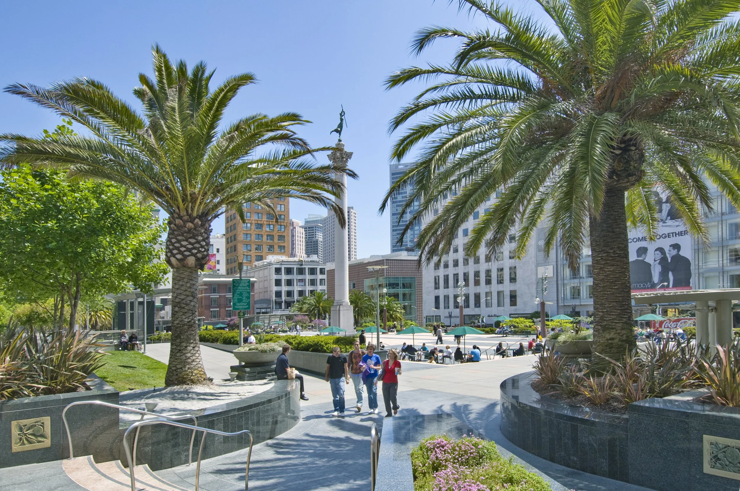 City park with palm trees, people walking, sitting, and socializing, surrounded by tall buildings and a statue on a sunny day.