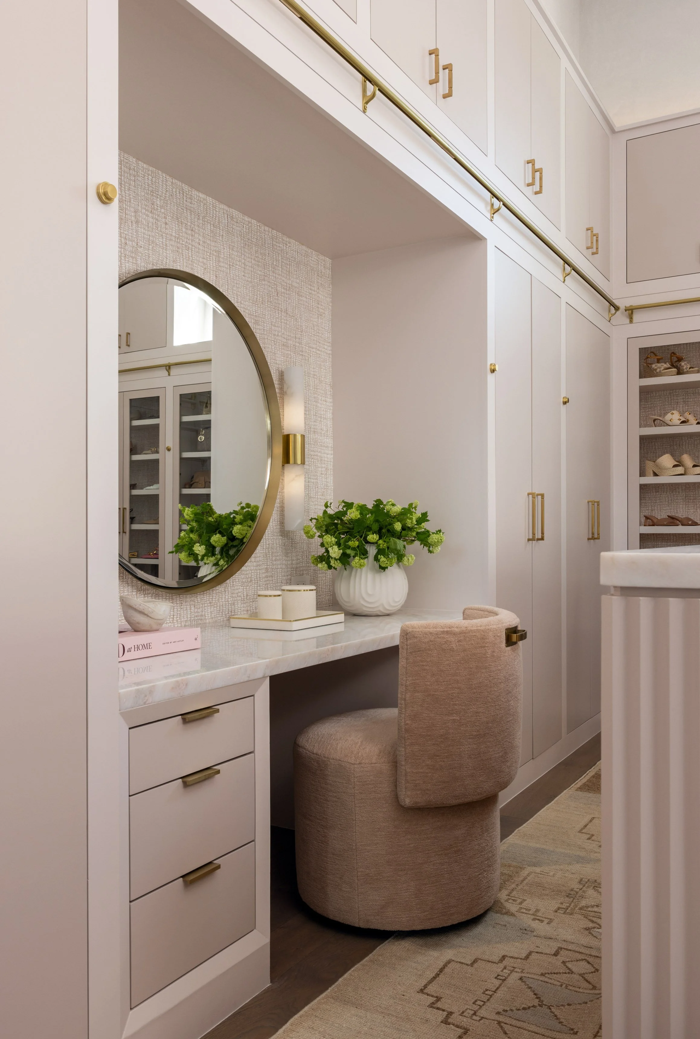 A cozy vanity area with a round mirror, a white marble top desk, a beige upholstered chair, decorative green flowers in a white vase, and built-in white cabinets with gold handles, complete with a textured beige wall and a small rug on the wooden flo