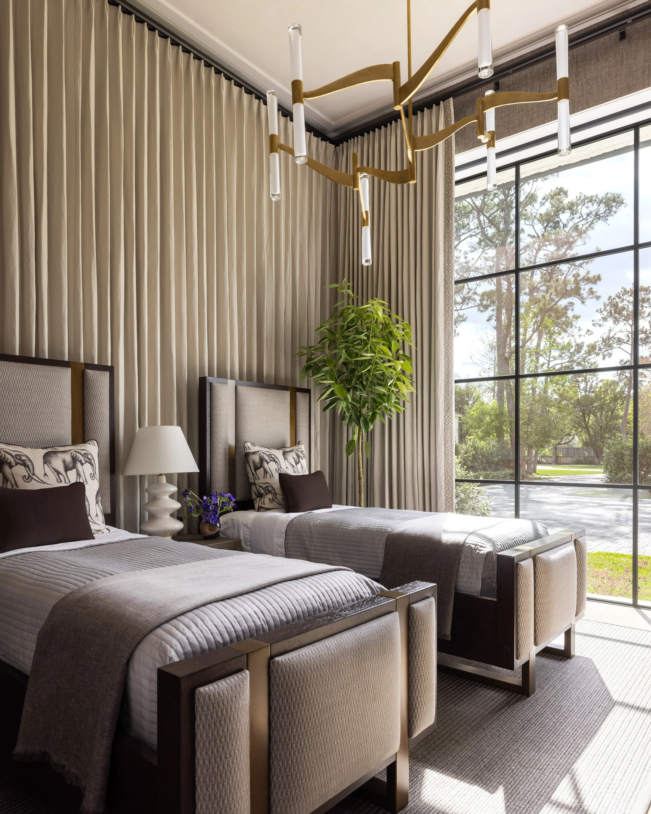 Benjamin Johnston Design guest bedroom with twin upholstered beds, elephant print pillows, brass chandelier, floor to ceiling drapery, upholstered bench, black steel windows with garden views, and potted indoor tree.