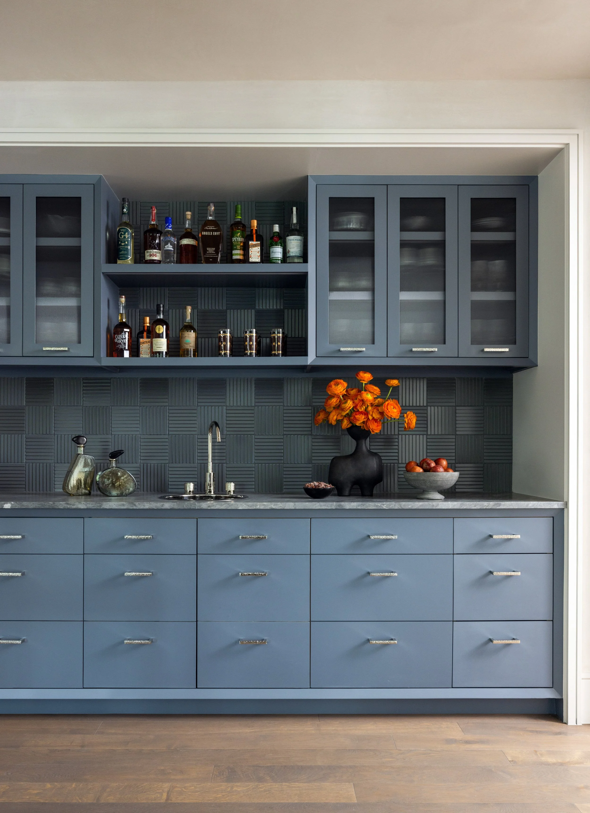 Blue kitchen cabinets with glass-front doors, a countertop with a black vase of orange flowers, bowl of apples, two glass bottles, and a black faucet under a gray tiled backsplash.