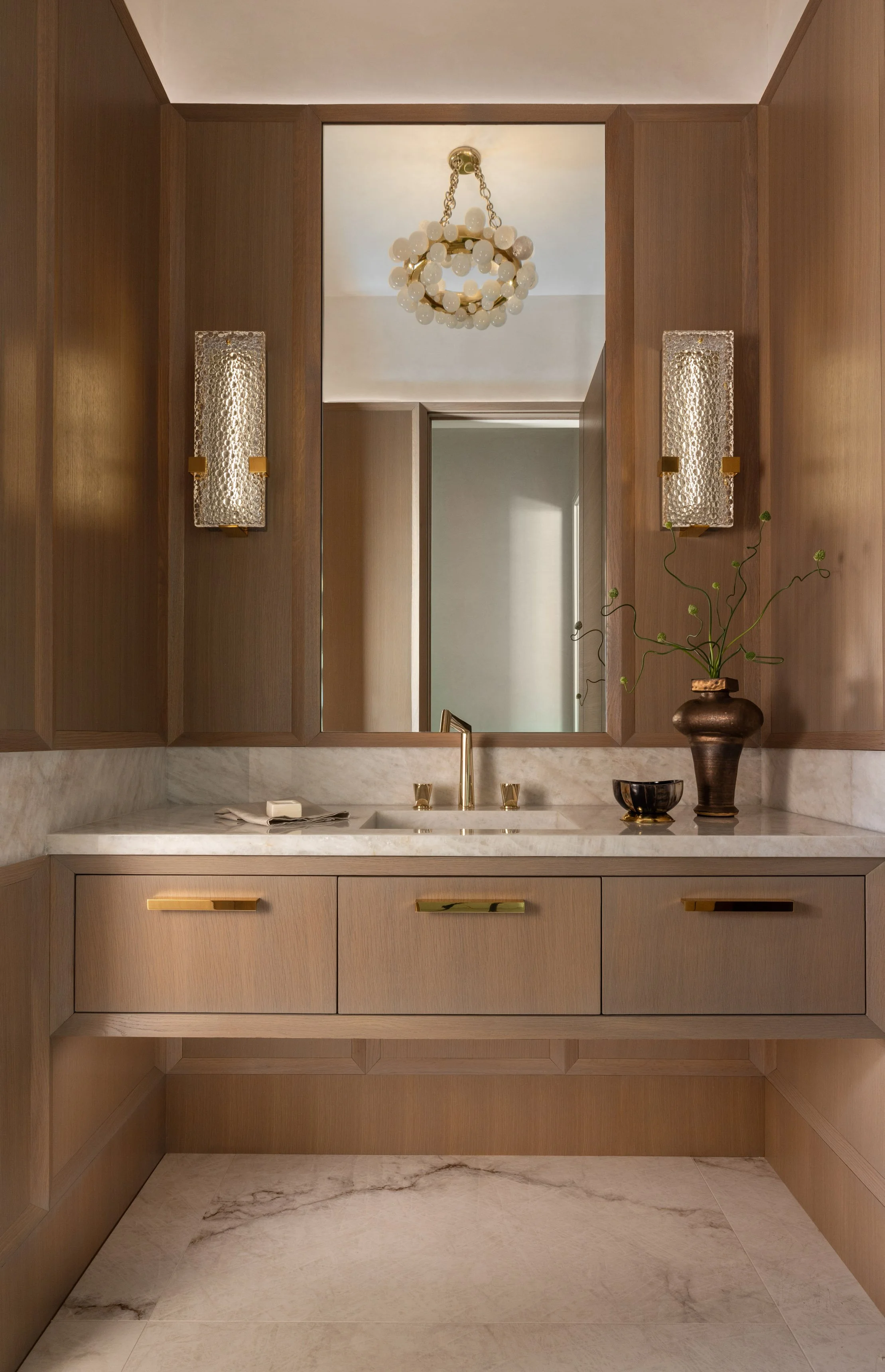 Bathroom vanity with wooden cabinetry, a white marble countertop, a mirror, a faucet, gold accents, decorative wall sconces, a potted plant, and a bowl.