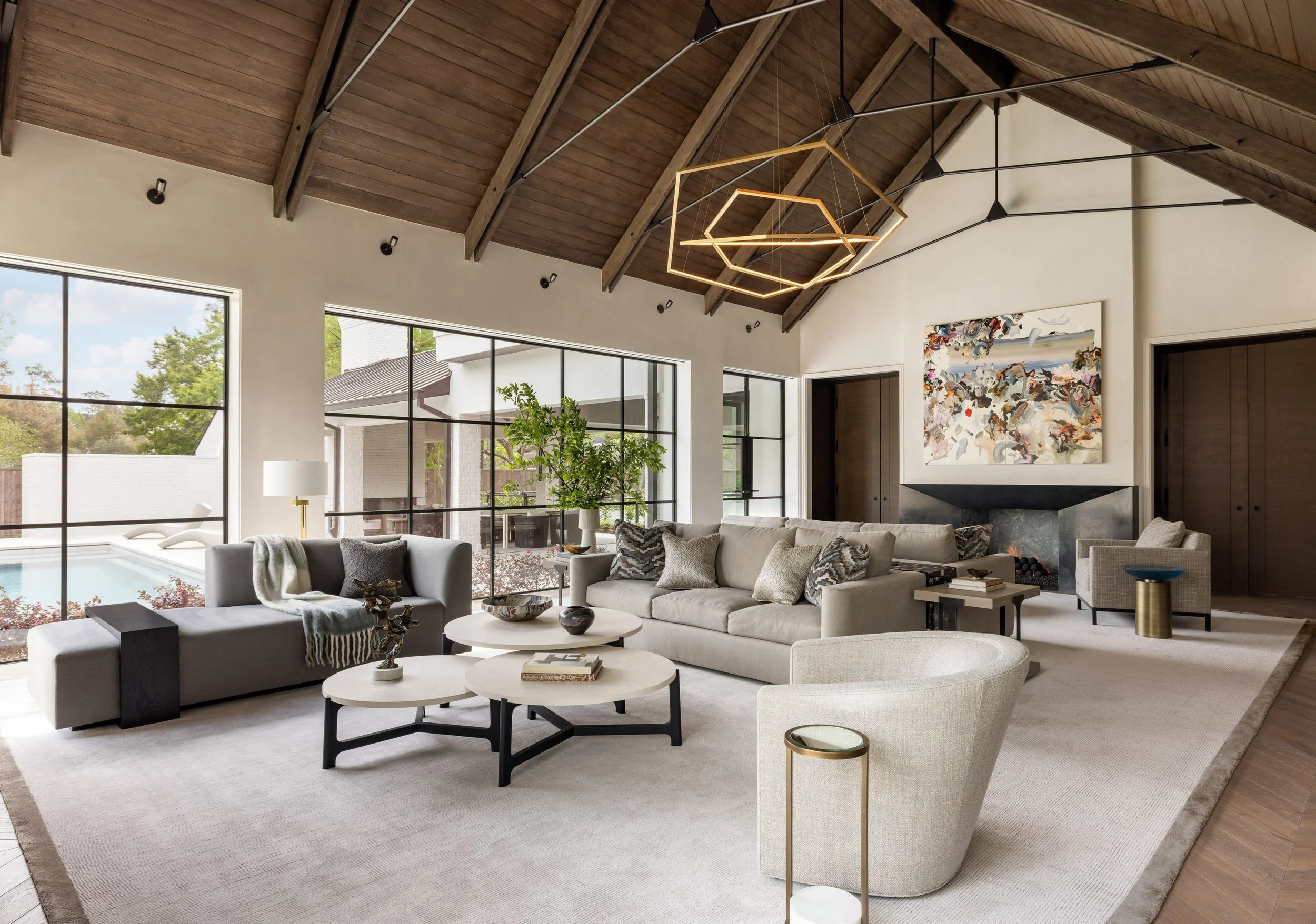 Benjamin Johnston Design great room wide angle view featuring vaulted wood ceiling with exposed beams, geometric brass chandelier, curved lounge chair, tiered round coffee tables, and neutral sectional sofas, pool views through steel windows.