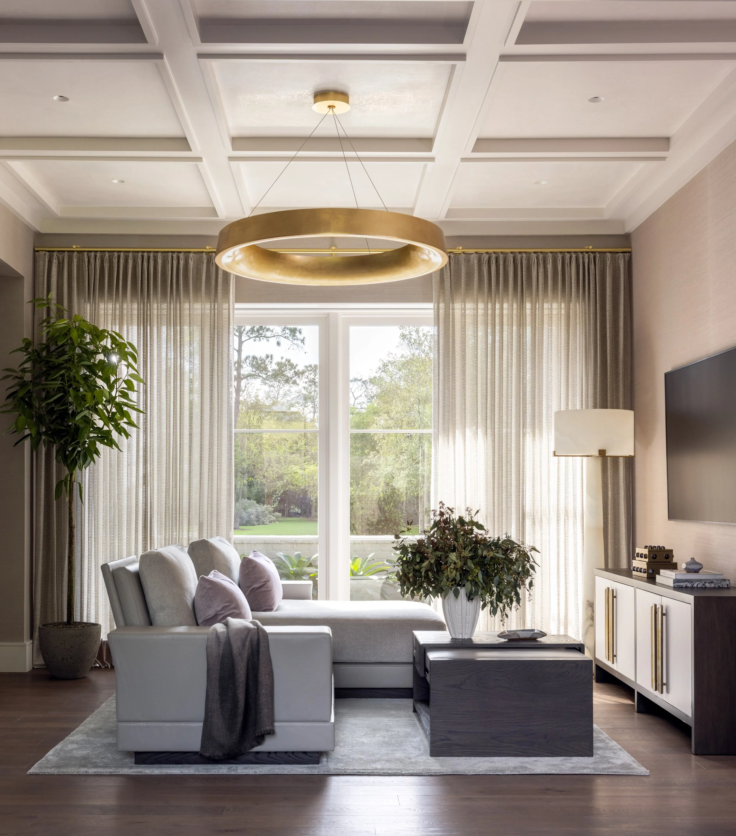 Benjamin Johnston Design Houston primary suite featuring coffered ceiling, oversized brass ring chandelier, gray sectional sofa, dark wood coffee table, sheer floor-to-ceiling drapery, and garden views.