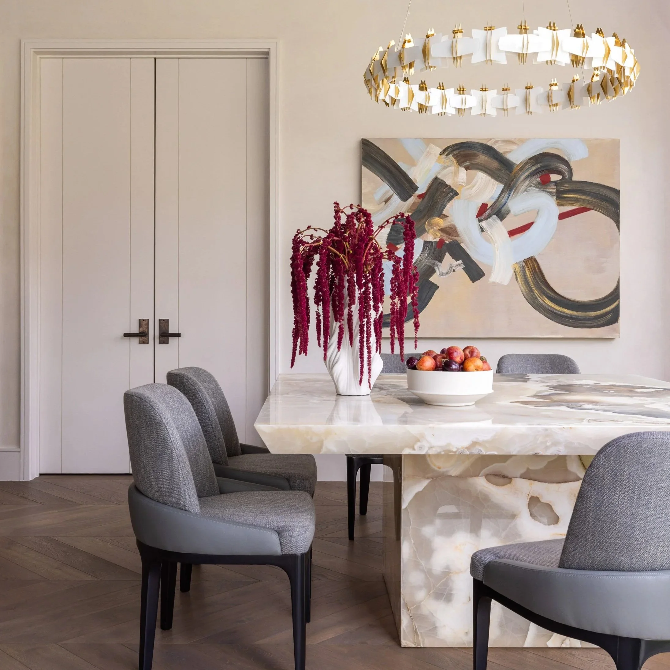 Benjamin Johnston Design Houston dining room detail featuring sculptural onyx dining table, gray upholstered dining chairs, tiered brass chandelier, bold abstract artwork, and dramatic amaranthus floral arrangement.