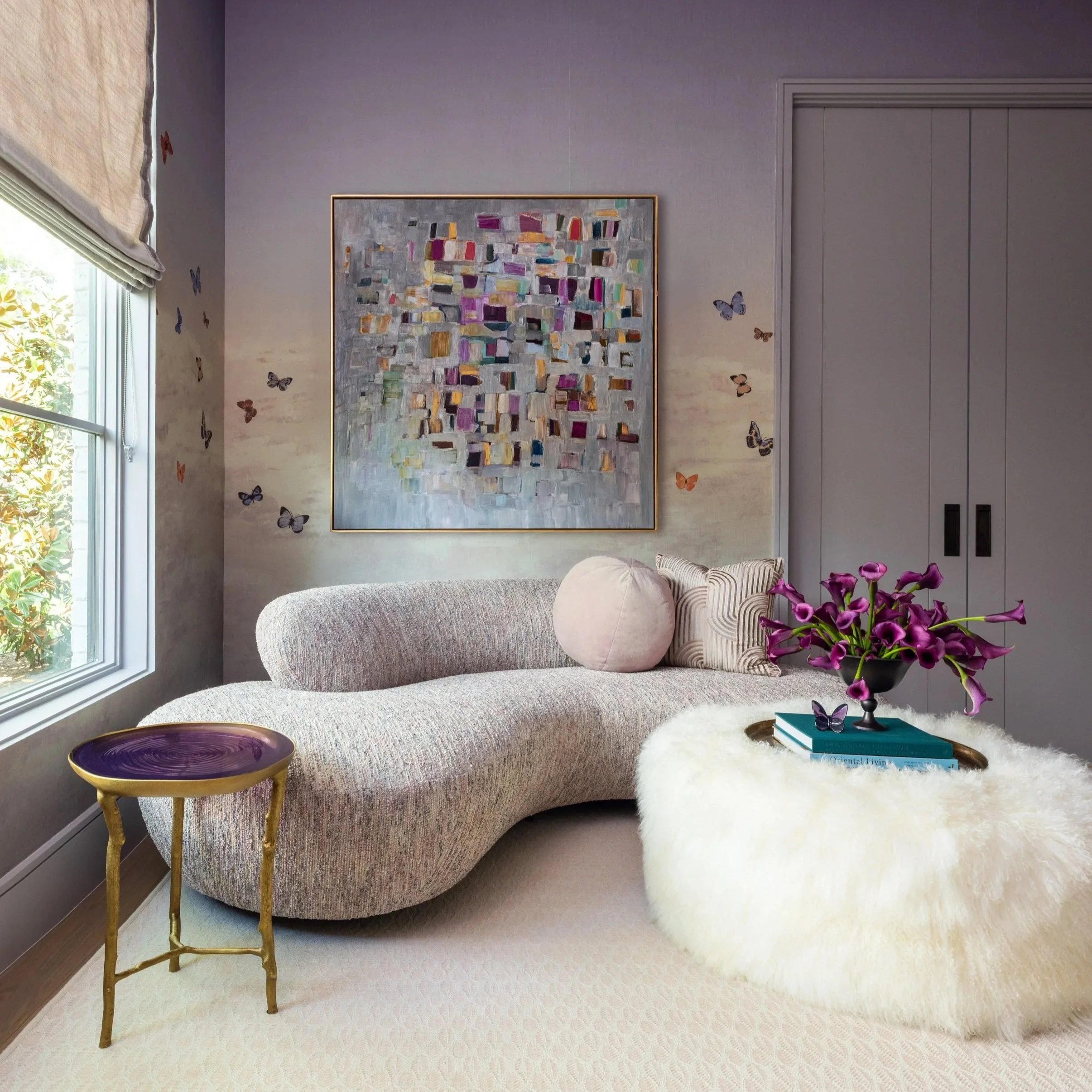 Benjamin Johnston Design children's lounge featuring lavender walls with butterfly wallpaper, curved boucle sofa, fluffy round ottoman, colorful abstract artwork, purple resin side table, and purple calla lily arrangement.