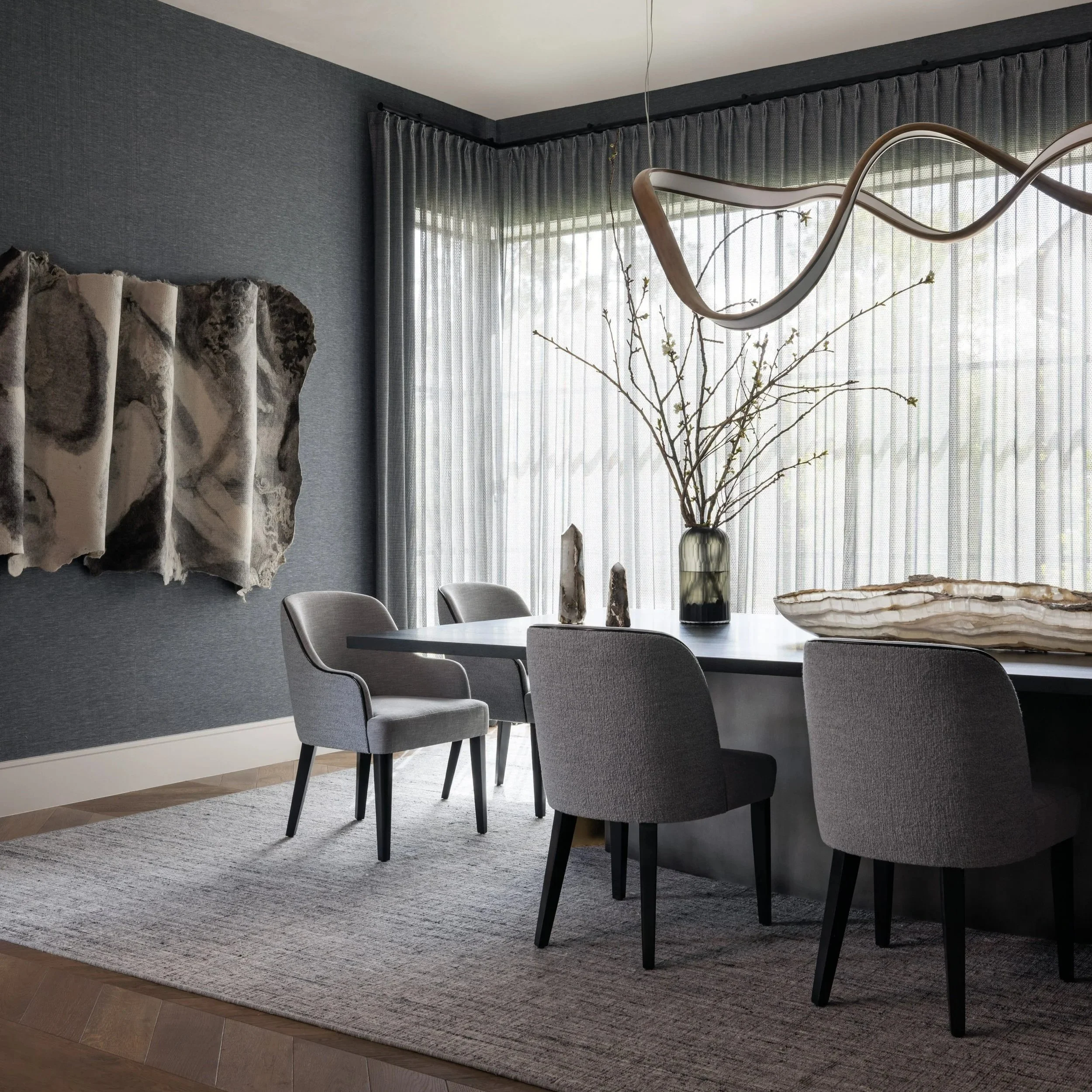 Benjamin Johnston Design Houston formal dining room featuring dark grasscloth walls, sculptural brass ribbon chandelier, gray upholstered dining chairs, petrified wood wall sculpture, crystal mineral centerpiece, and sheer floor-to-ceiling drapery.