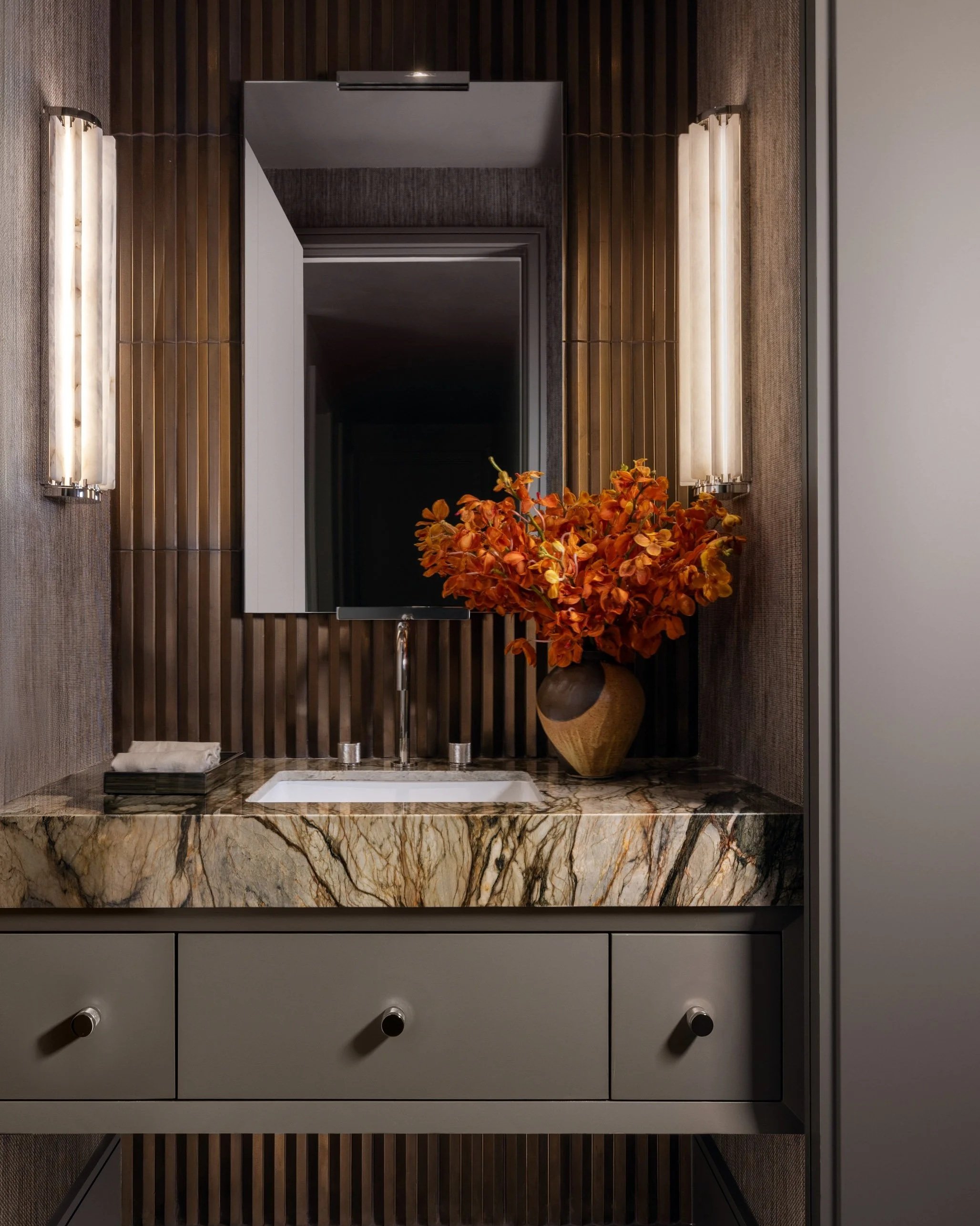 Benjamin Johnston Design powder room featuring dramatic veined marble countertop, fluted dark wood accent wall, alabaster tube sconces, floating vanity with sage cabinetry, and oversized orange orchid arrangement.