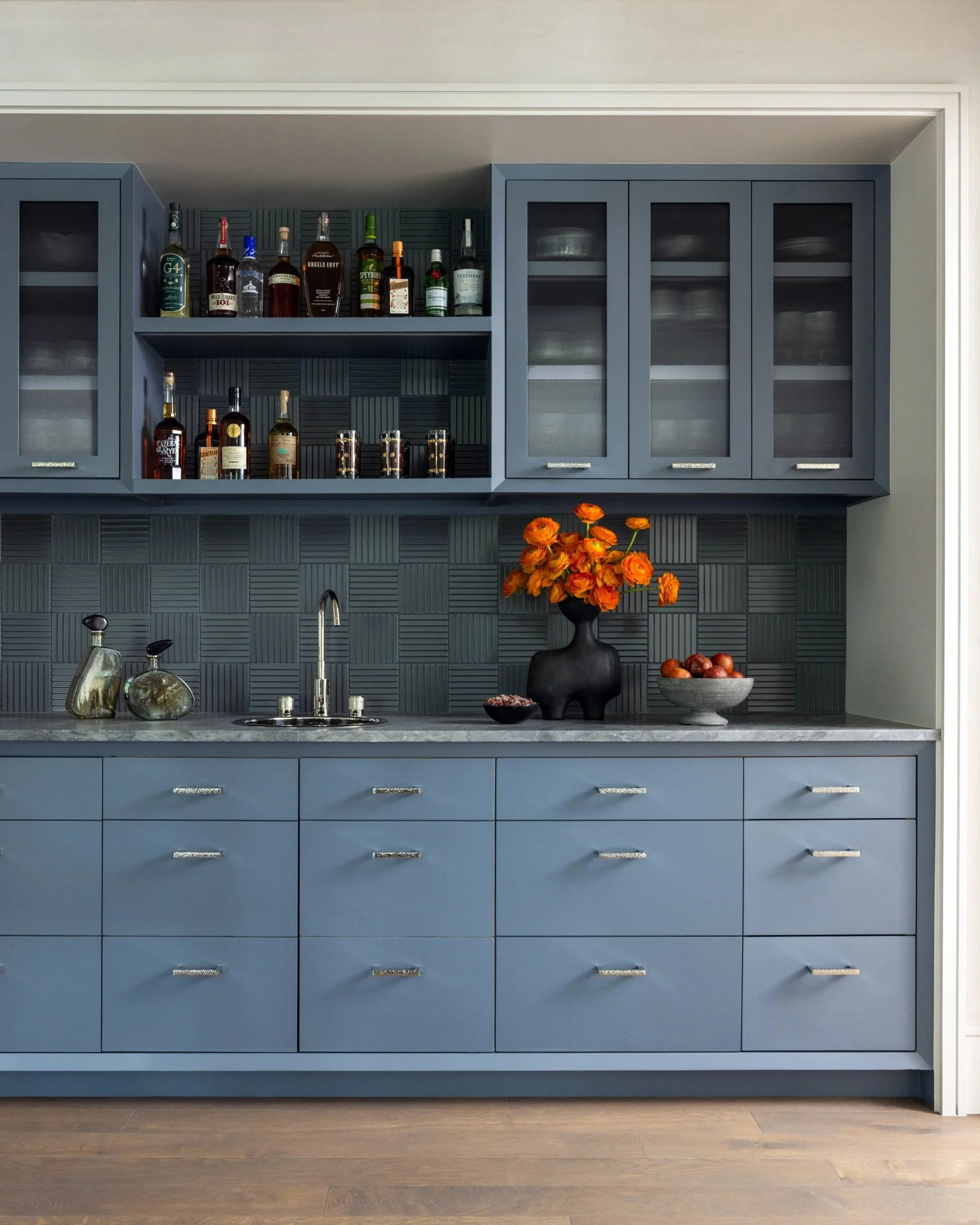 Benjamin Johnston Design home bar featuring slate blue cabinetry with nickel hardware, dimensional basketweave tile backsplash, marble countertop, open spirit shelving, and orange ranunculus floral arrangement.