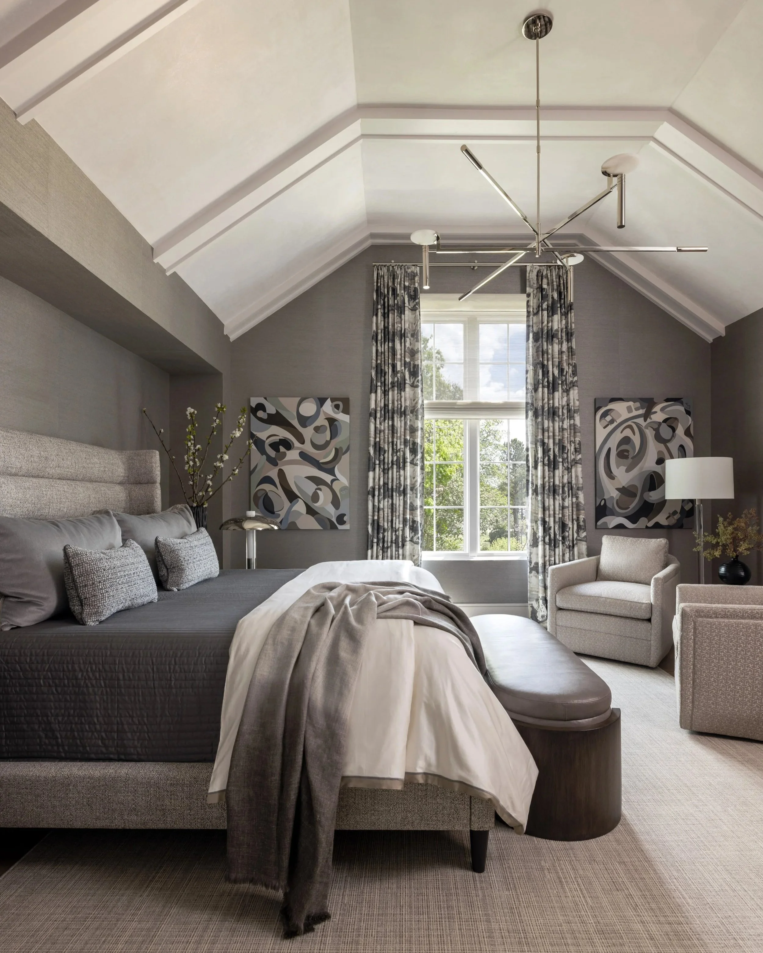 Benjamin Johnston Design primary bedroom featuring vaulted ceiling with architectural molding, polished nickel sputnik chandelier, upholstered channel tufted headboard, gray grasscloth walls, abstract artwork, floral drapery, and sitting area.