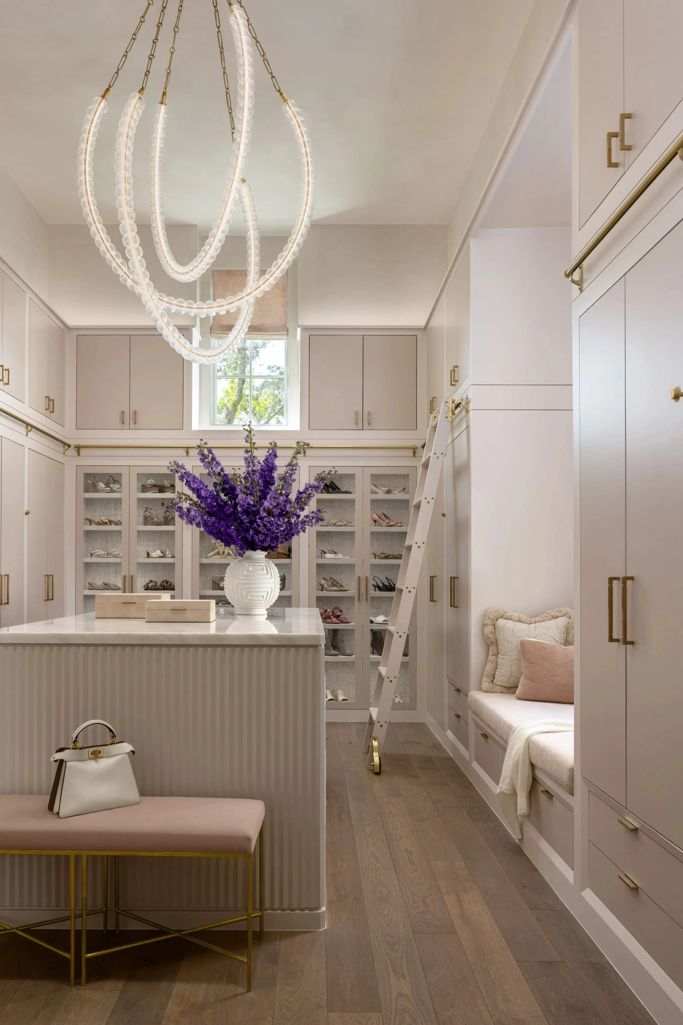 Benjamin Johnston Design primary closet featuring blush cabinetry with brass hardware, glass front shoe display cabinets, brass library ladder, crystal chandelier, fluted island with marble countertop, and built-in window seat.