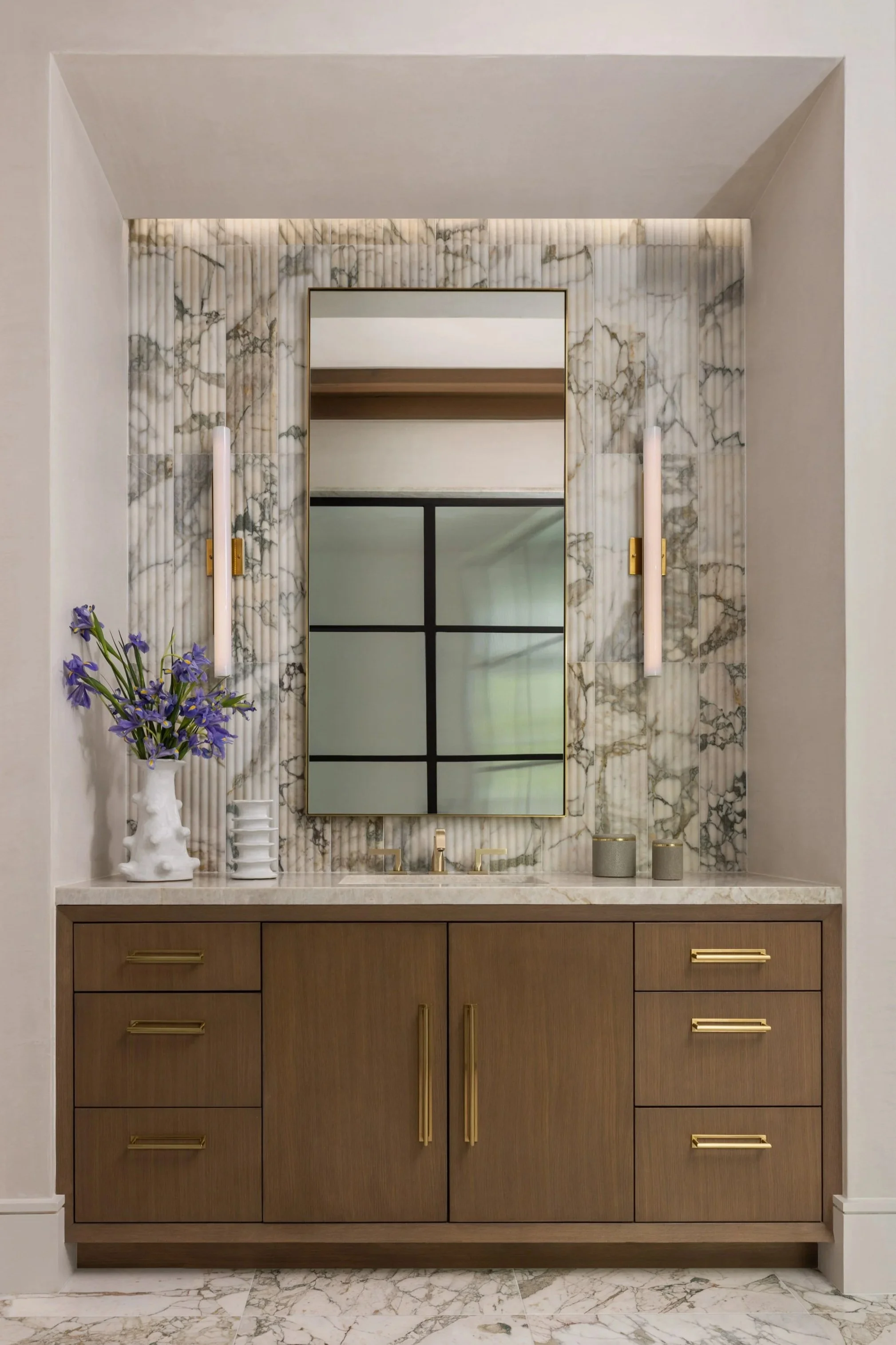 Benjamin Johnston Design primary bathroom vanity featuring fluted marble slab feature wall, walnut cabinetry with brass hardware, brass tube sconces, framed mirror, marble countertop, and marble tile floor.