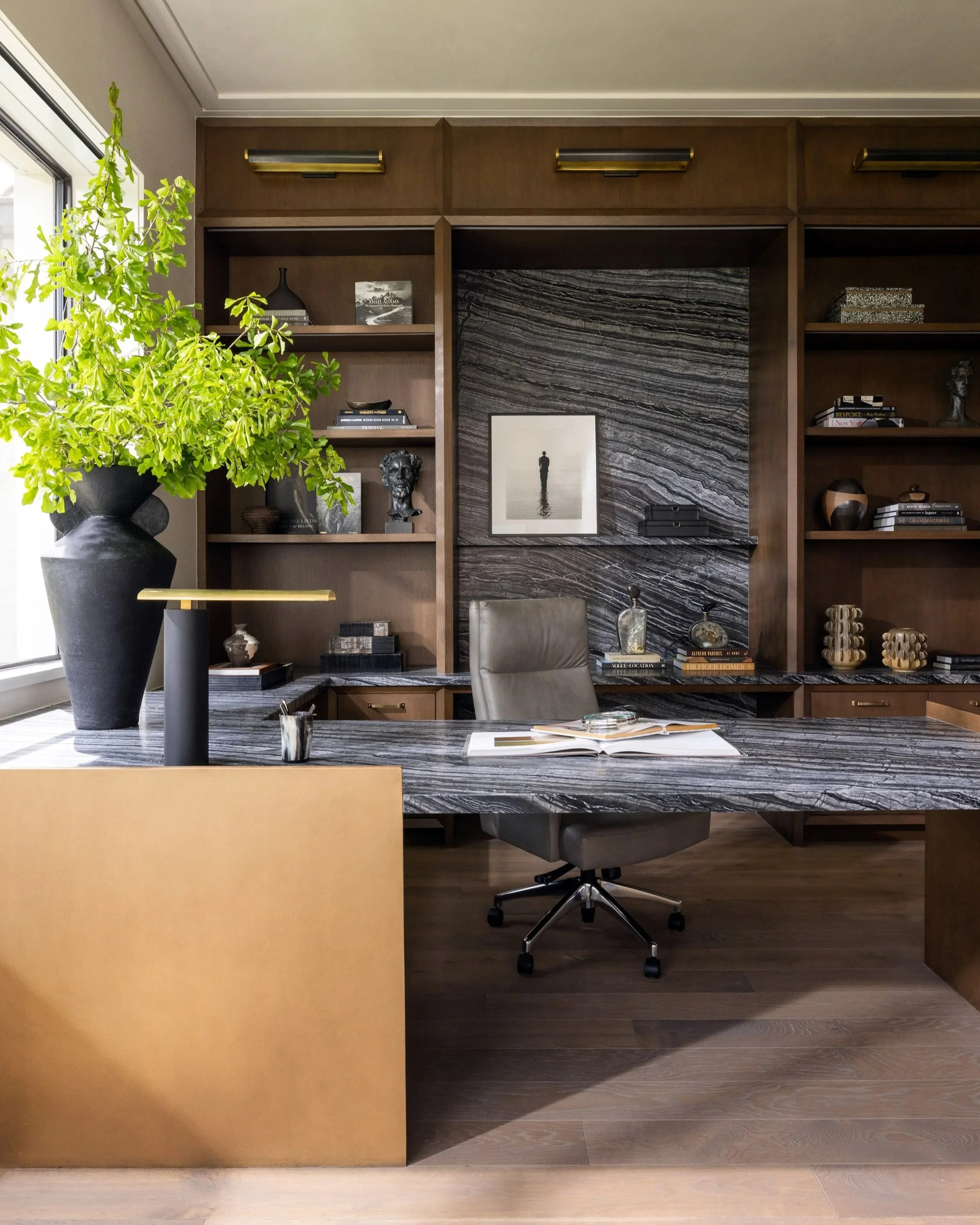 Benjamin Johnston Design Houston home office featuring veined marble desk, walnut built-in shelving with brass accents, dramatic marble slab feature wall, leather task chair, curated art and sculpture, and large botanical arrangement.