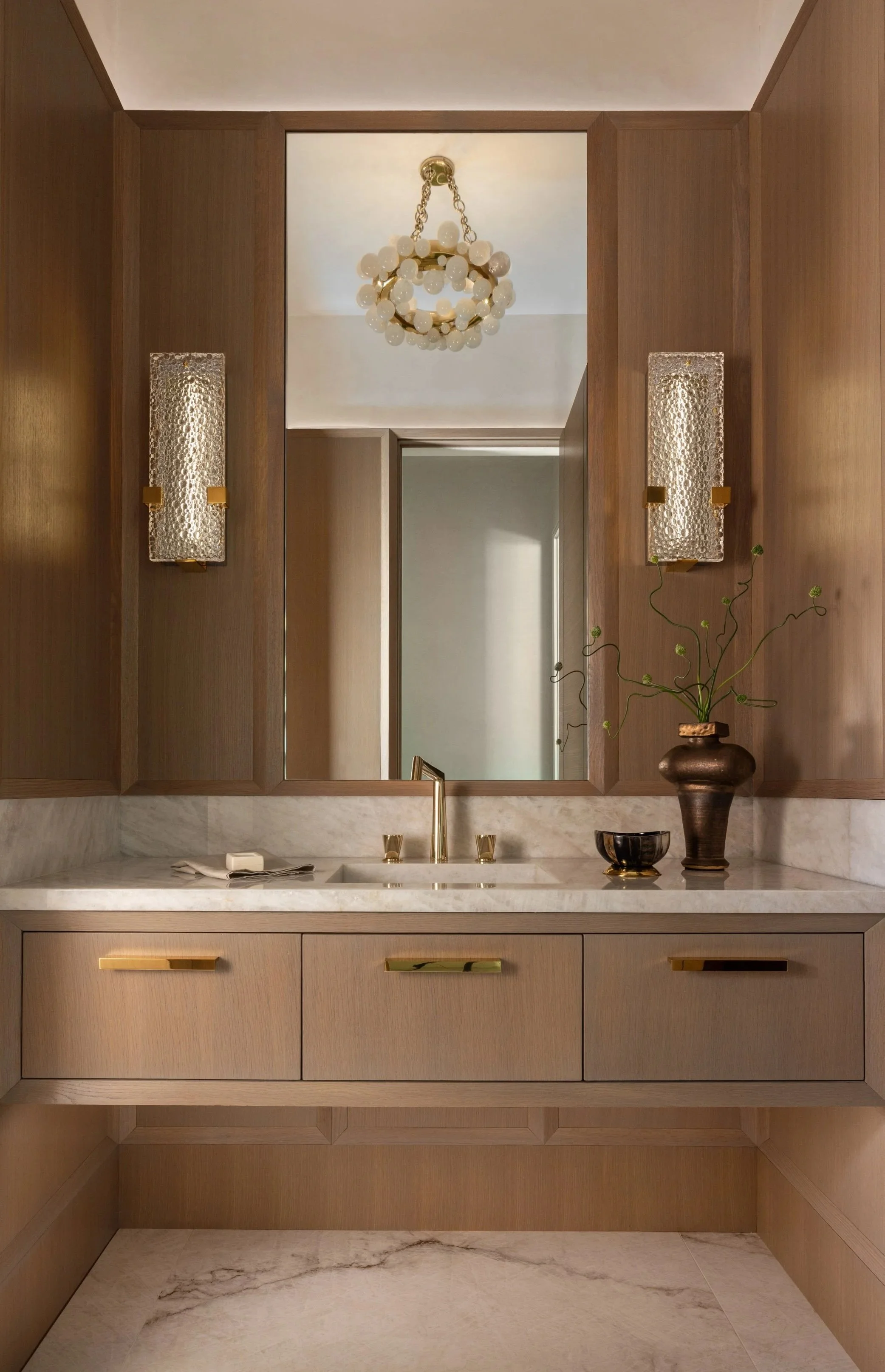 Benjamin Johnston Design Houston powder room featuring warm oak paneled walls, floating vanity with marble countertop, brass hardware, crystal wall sconces, and globe chandelier reflected in mirror.