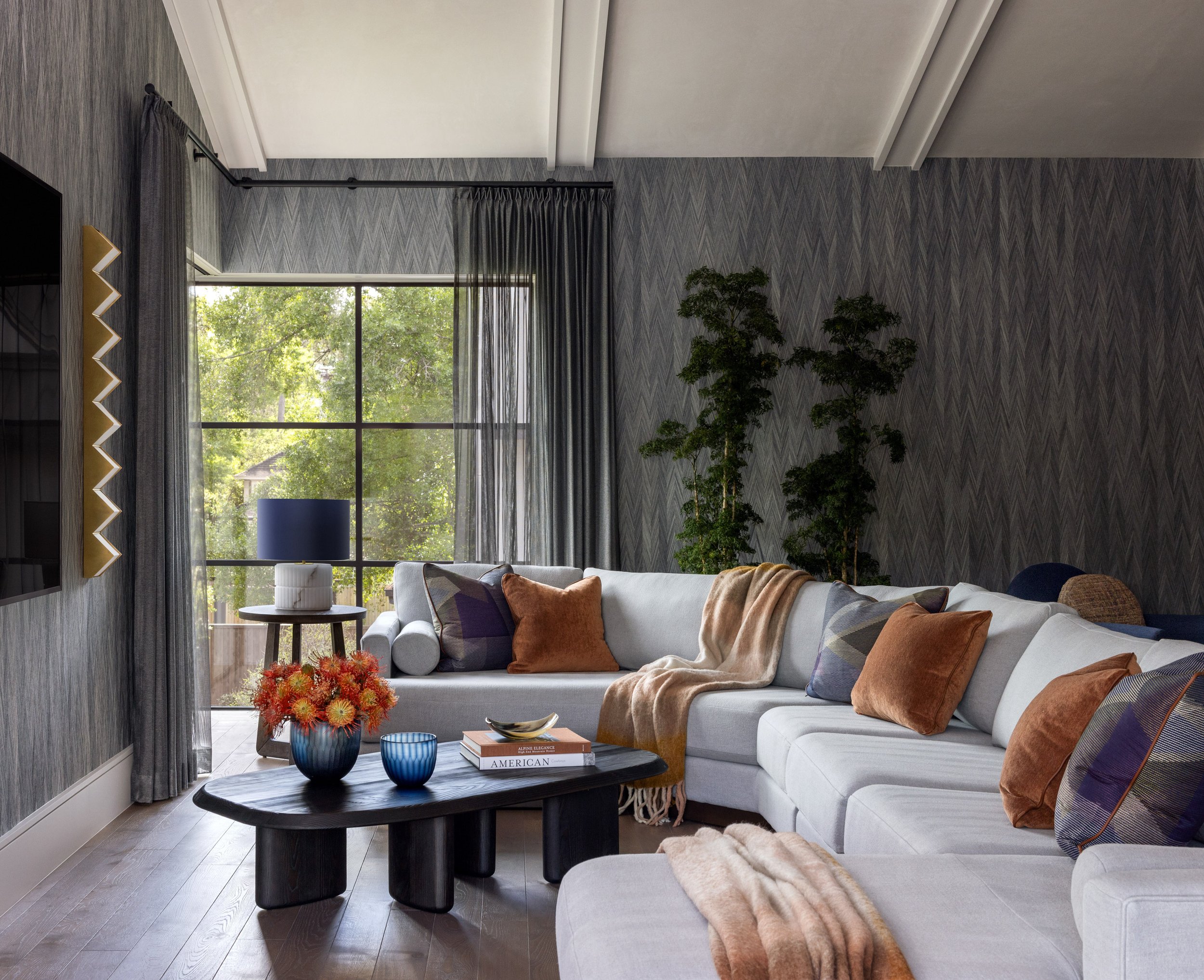 A cozy living room with a large white sectional sofa decorated with orange, purple, and plaid pillows, a dark round coffee table with blue glassware and books, a tall green indoor plant near a window with gray curtains, and a wall with textured gray 